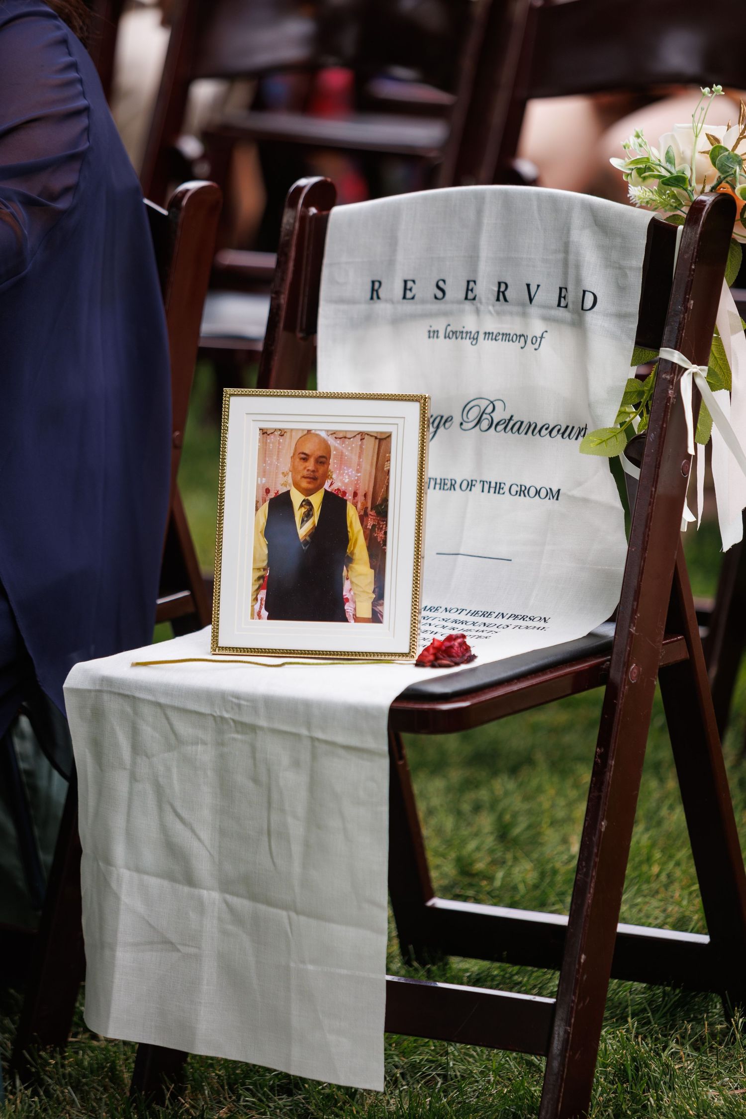 Reserved memorial chair with framed photo honoring Jorge Betancourt, Father of the Groom, at an outdoor wedding ceremony.