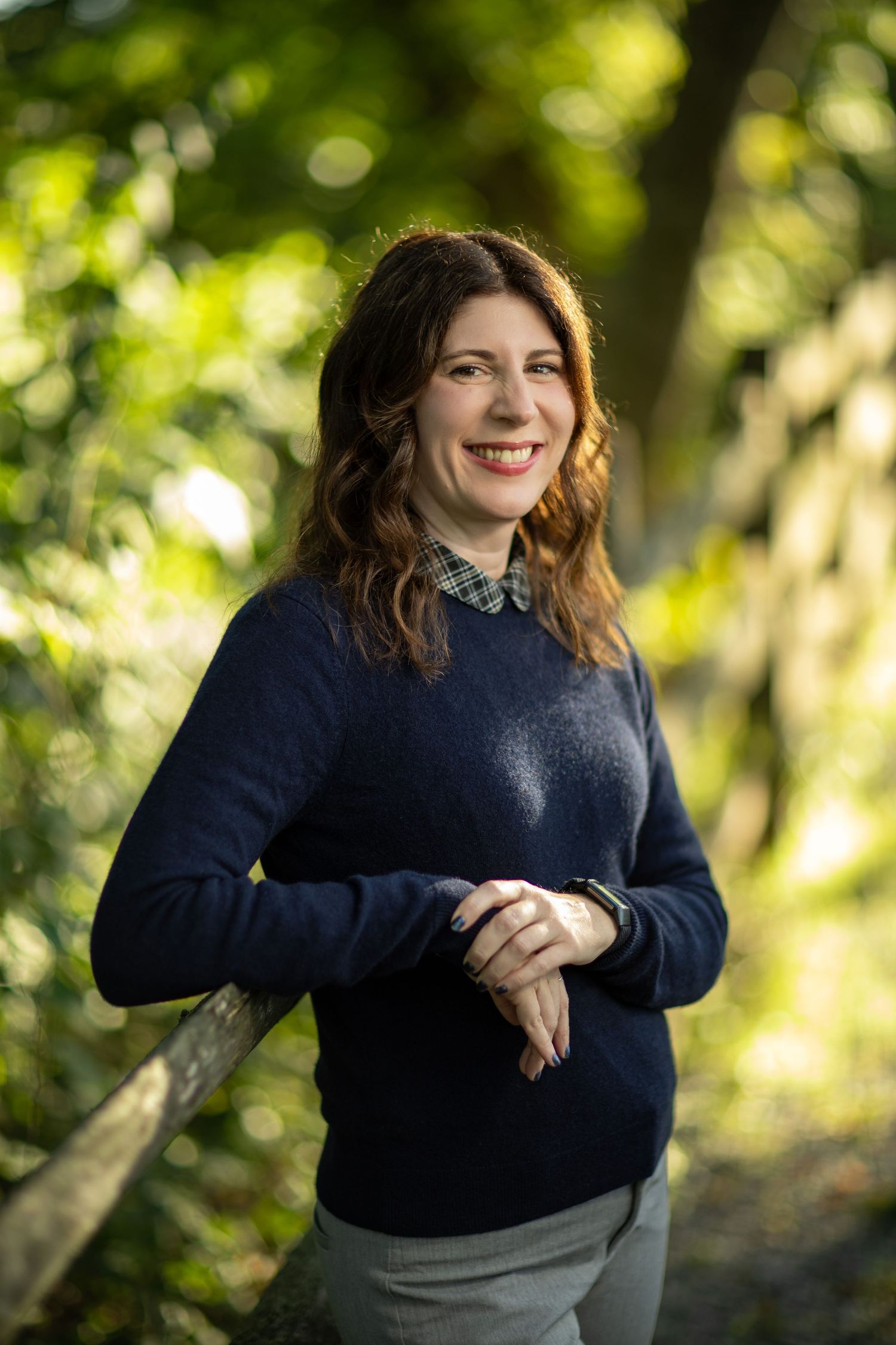 A person wearing a navy sweater poses against a blurred green forest background in natural lighting.