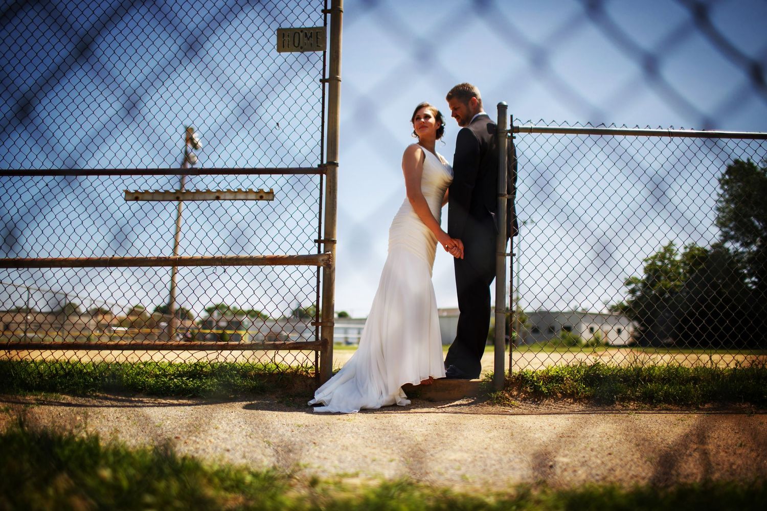 Wedding couple poses against chain link fence with cross and sunlight creating dramatic shadows.