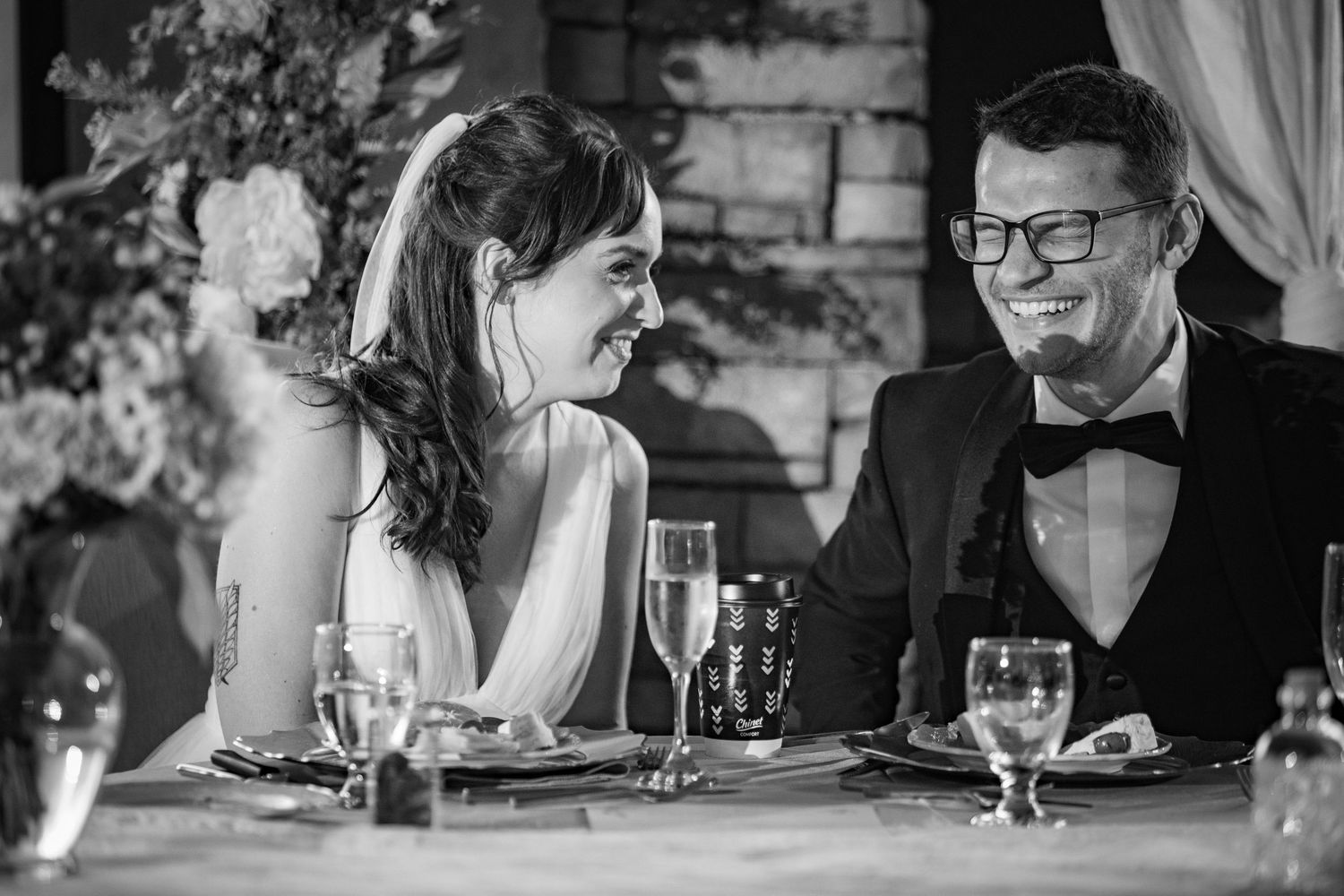 Bride and groom laugh together at wedding reception dinner table in black and white photo.
