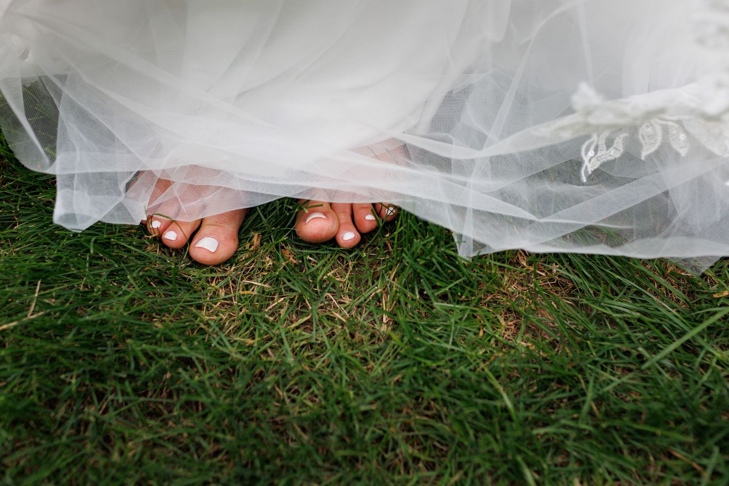 Close-up of bride's bare feet with painted toenails peeking beneath white tulle wedding dress on grass.