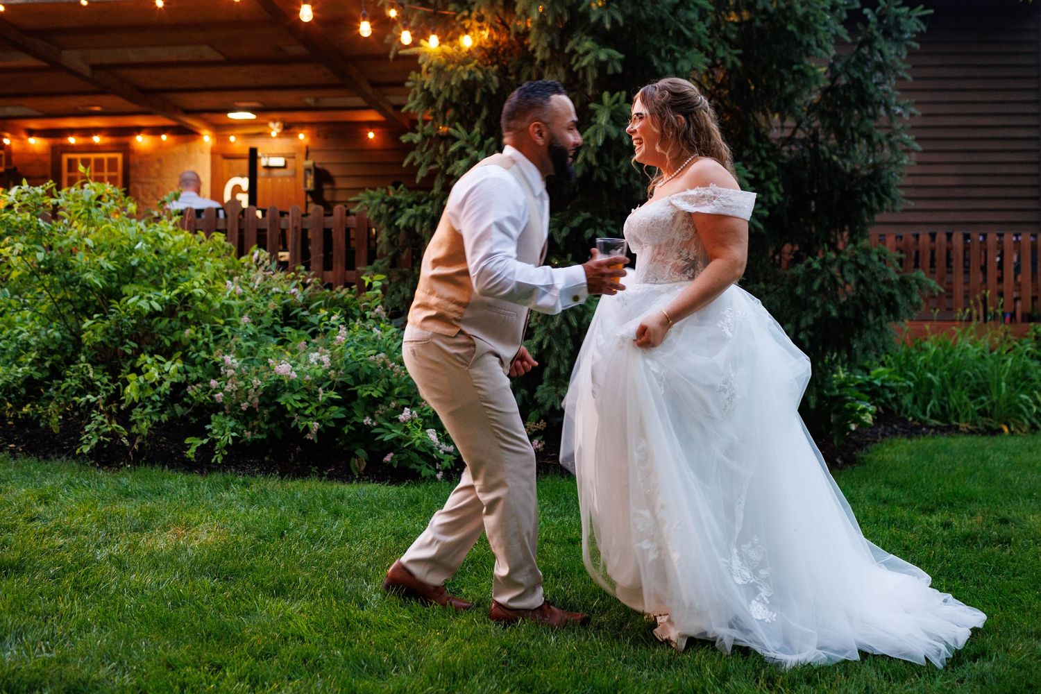Bride and groom dancing together in a garden at dusk with string lights glowing in the background.