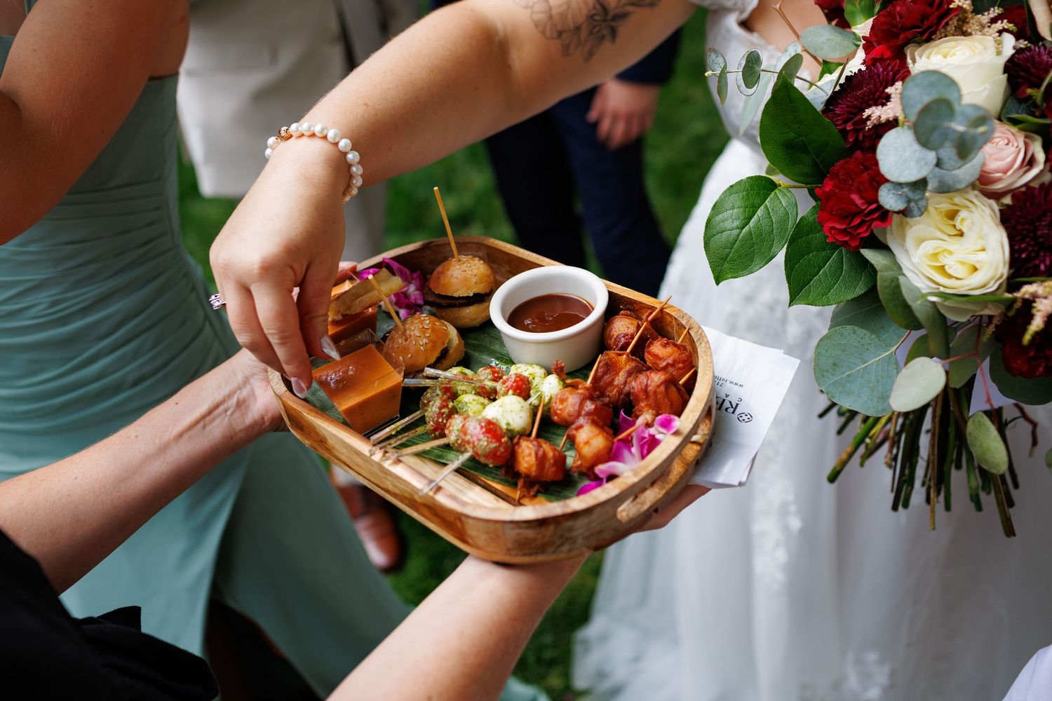 Wedding guests sharing a wooden tray of appetizers including sliders and skewers with dipping sauce.