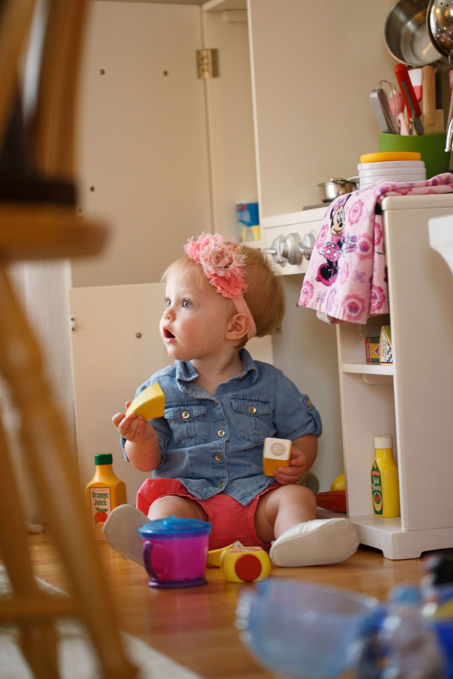 Baby in denim clothing sits among colorful toys in a casual indoor setting.