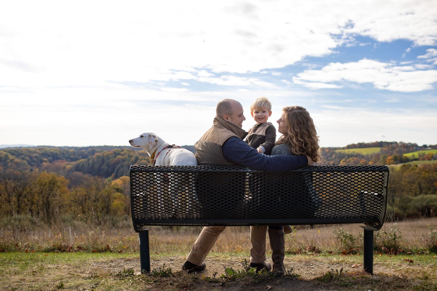 Three people sit together on a black metal bench overlooking an autumn mountain landscape on a sunny day.