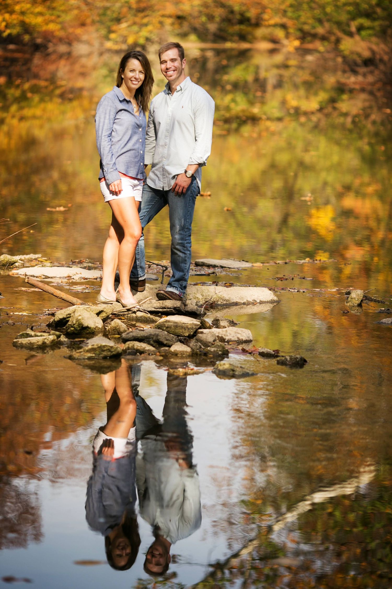 A couple stands on rocks by a calm pond with autumn foliage reflected in the water.