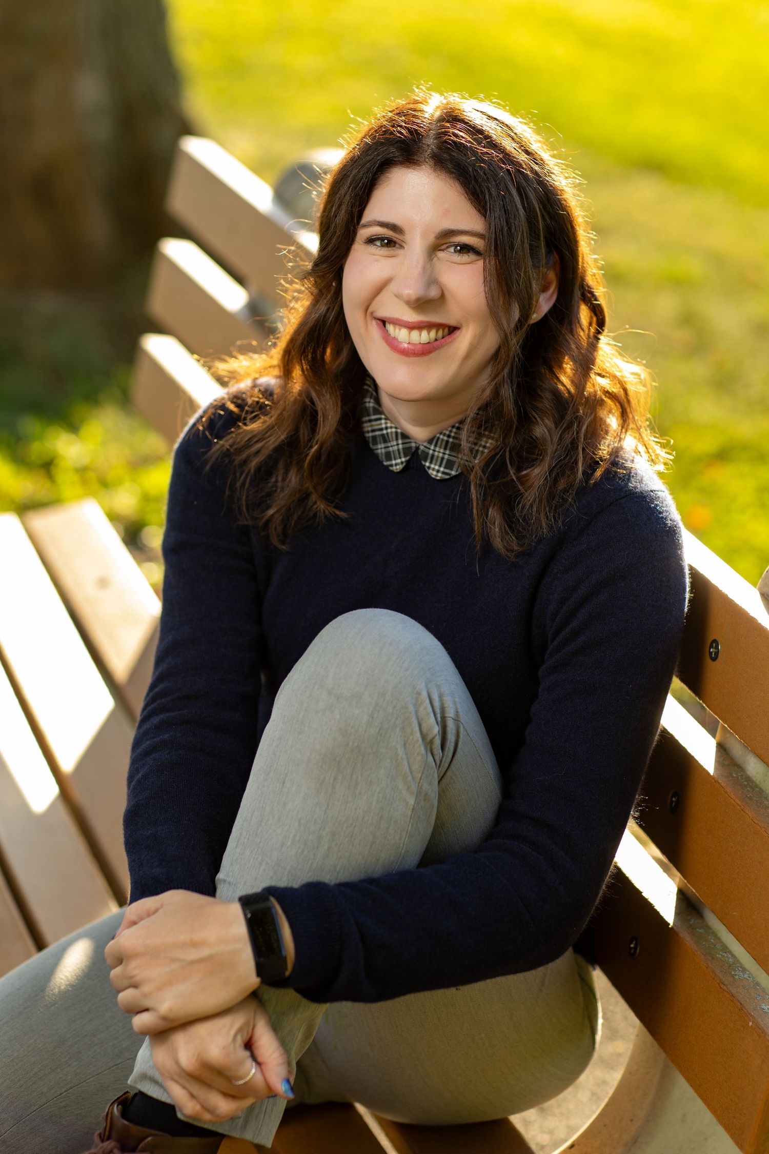 Woman in a black sweater sits on a wooden bench outdoors in warm sunlight during an autumn day.