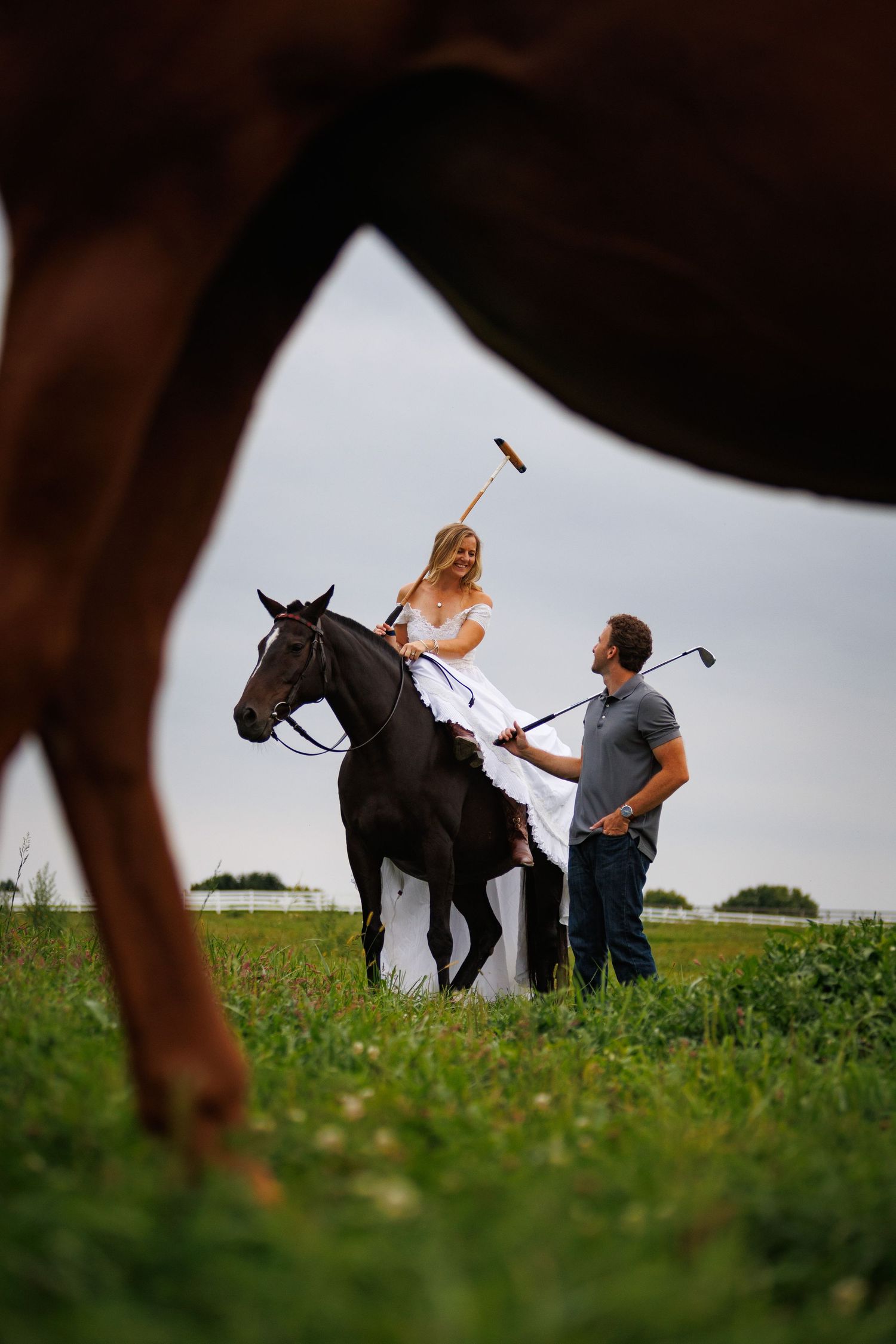 Woman in white dress on dark horse holds polo mallet while man stands beside her in green field.