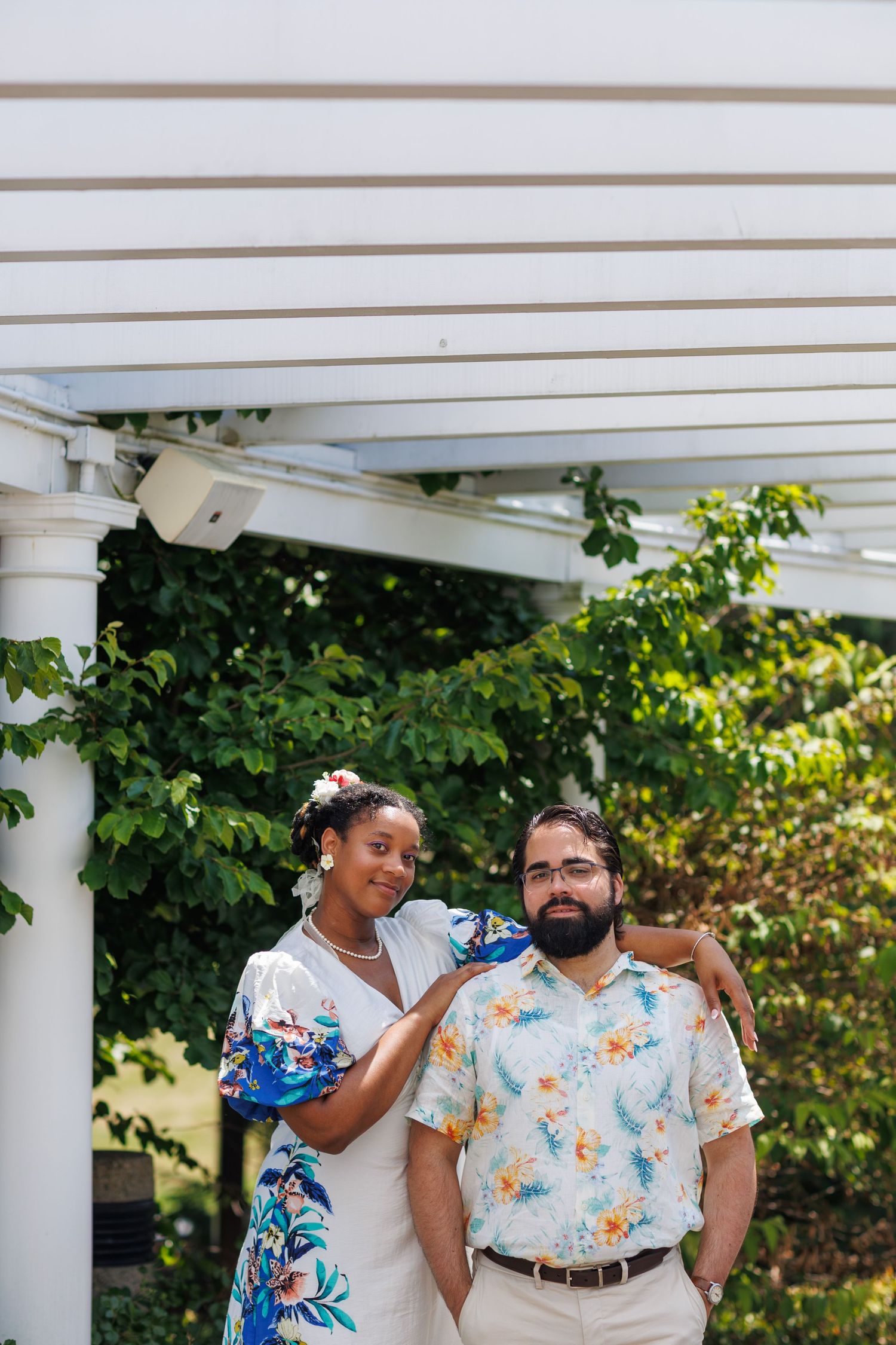 A couple poses under a white pergola surrounded by lush greenery, woman in floral dress rests hand on mans shoulder.
