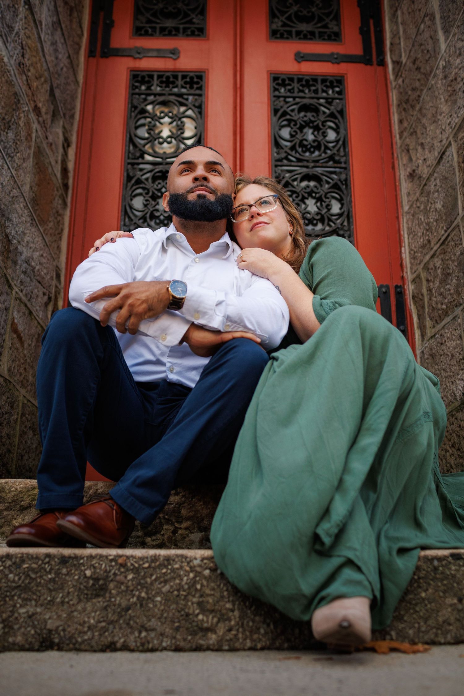 Romantic couple resting against red brick wall with ornate iron windows in intimate photo series.
