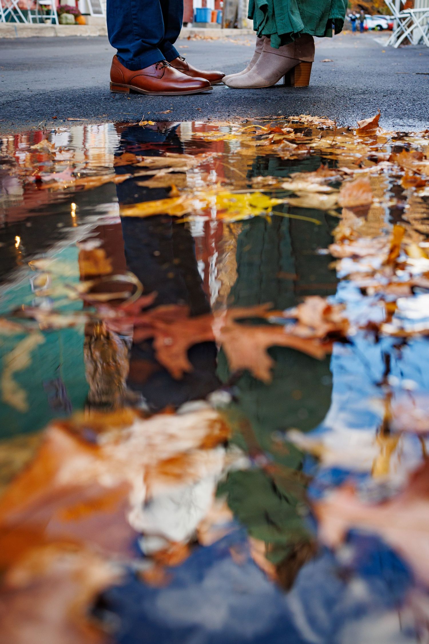 Autumn leaves reflect in a puddle creating a colorful mirror effect with people's feet visible at the edges.