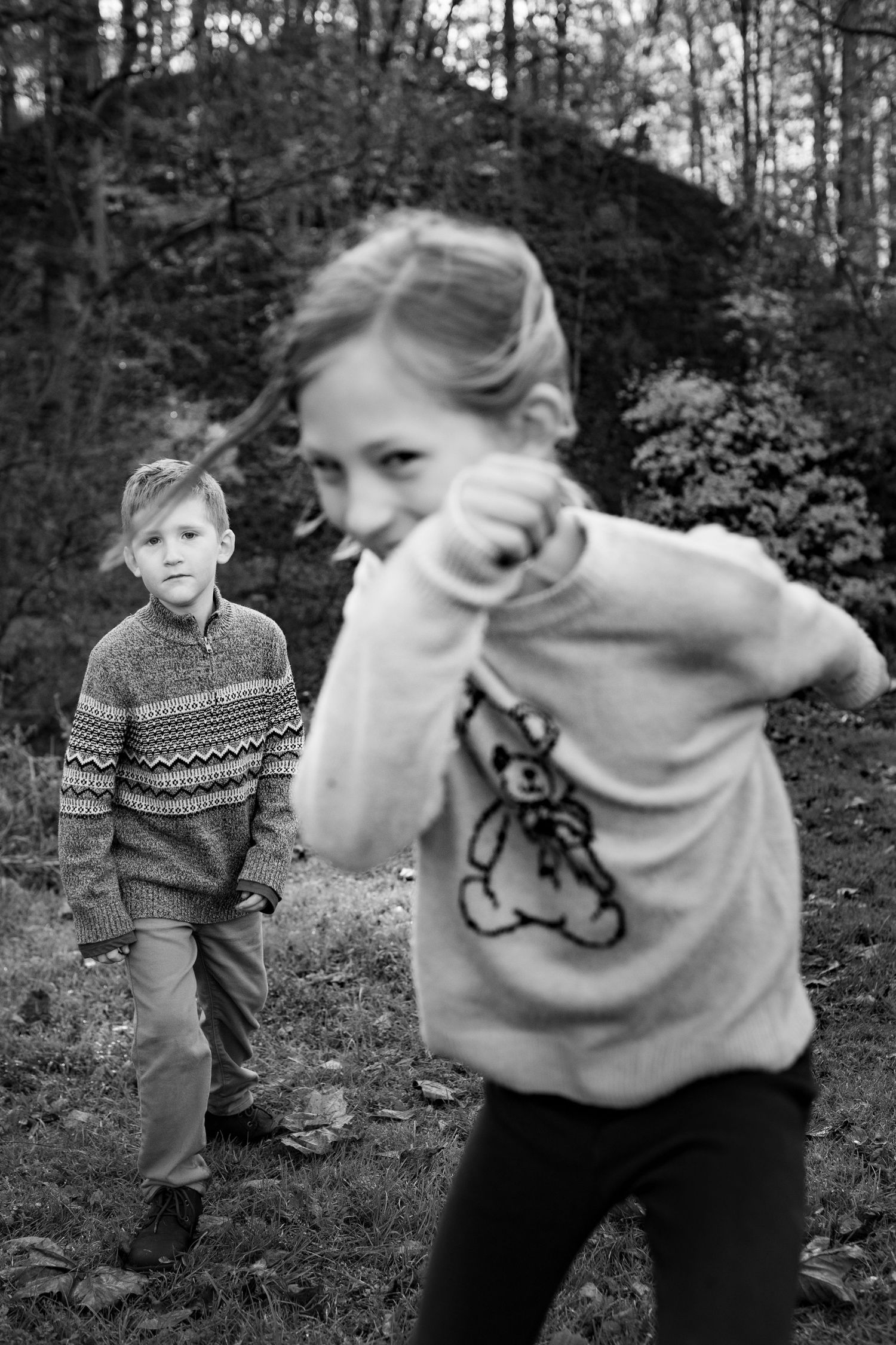 Two children in sweaters play outdoors in a black and white forest setting.