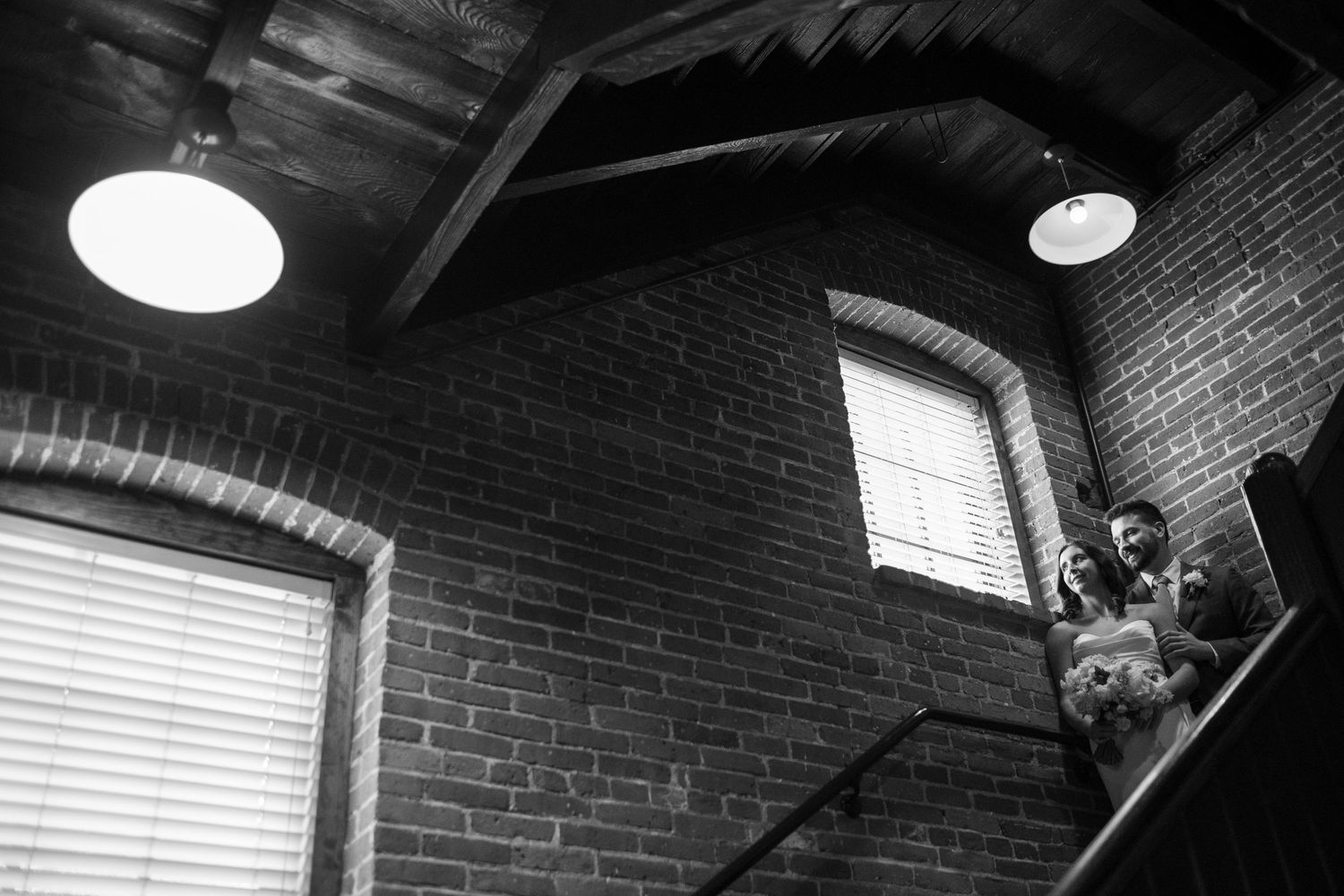 Black and white shot of couple descending staircase in rustic brick venue with pendant lights overhead.