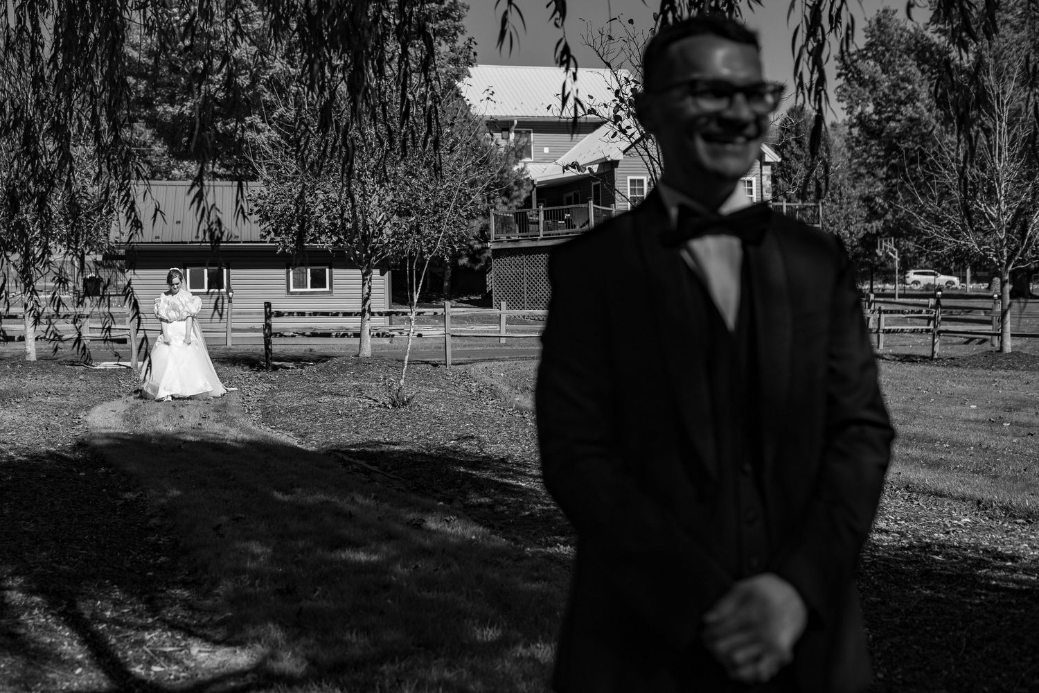 Black and white wedding photo of groom smiling in foreground with bride approaching across rustic farm venue.