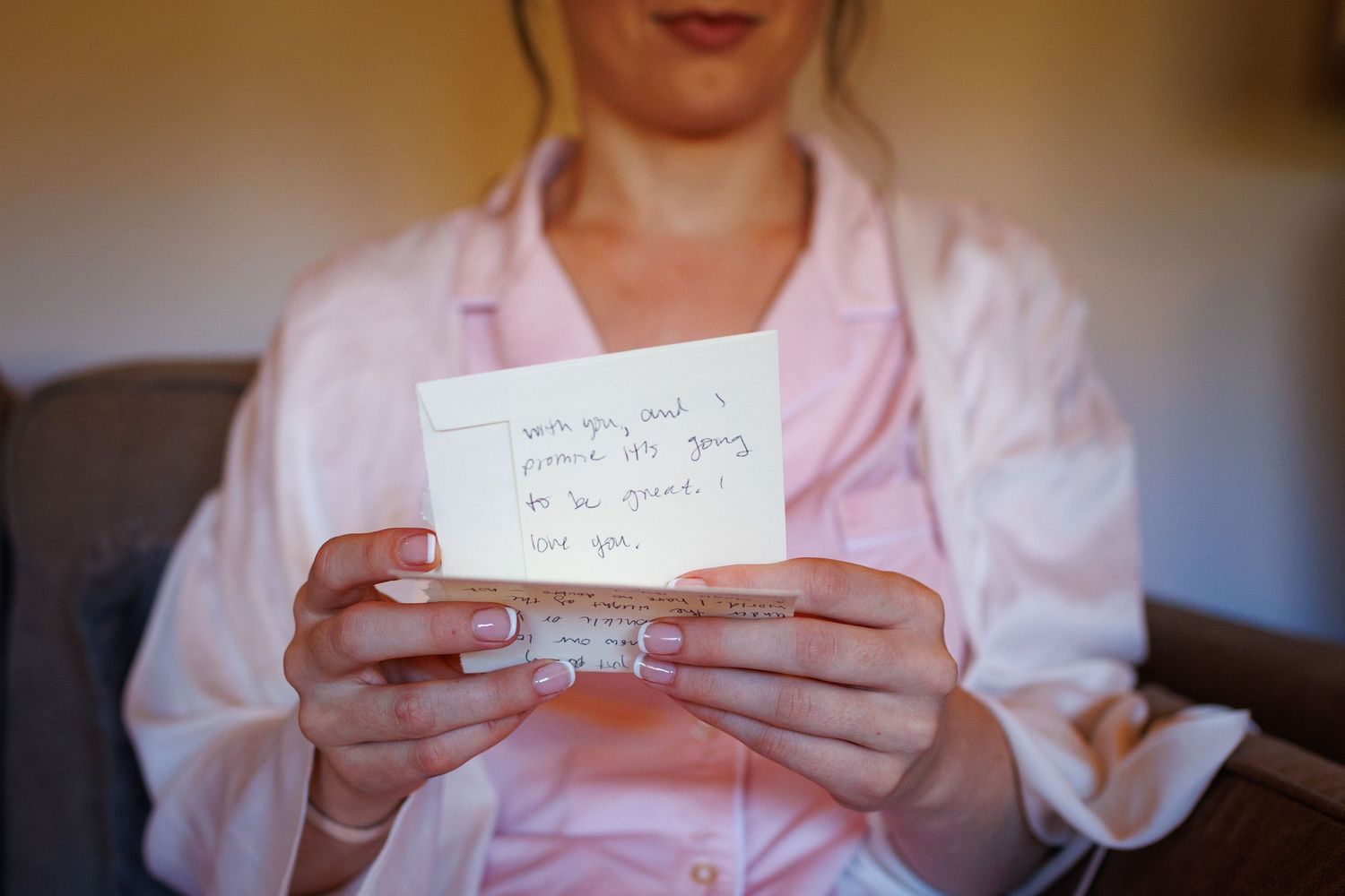 Bride in pink satin robe reads a heartfelt handwritten note card while holding it with manicured hands.
