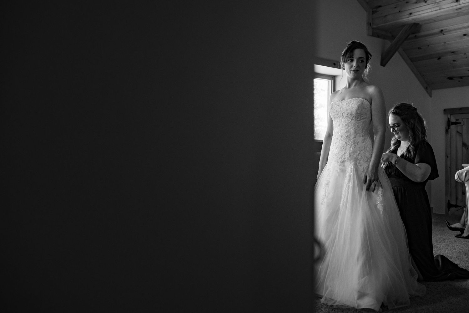 Black and white photo of a bride in a lace gown being helped by a bridesmaid near a window.