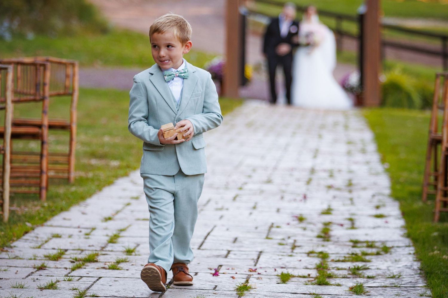 Young ring bearer in light blue suit walking down outdoor wedding ceremony aisle.