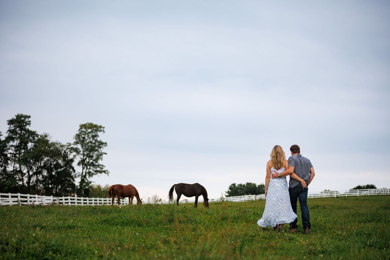 Bride and groom stand together watching horses graze on a lush green farm field with a white fence.