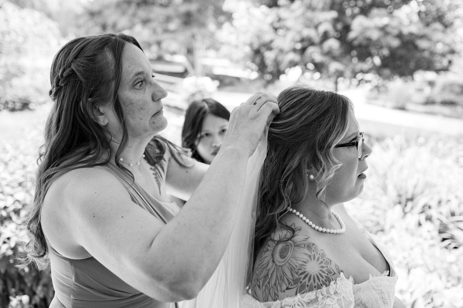 Black and white photo of a woman adjusting the hair of a bride wearing pearls outdoors.