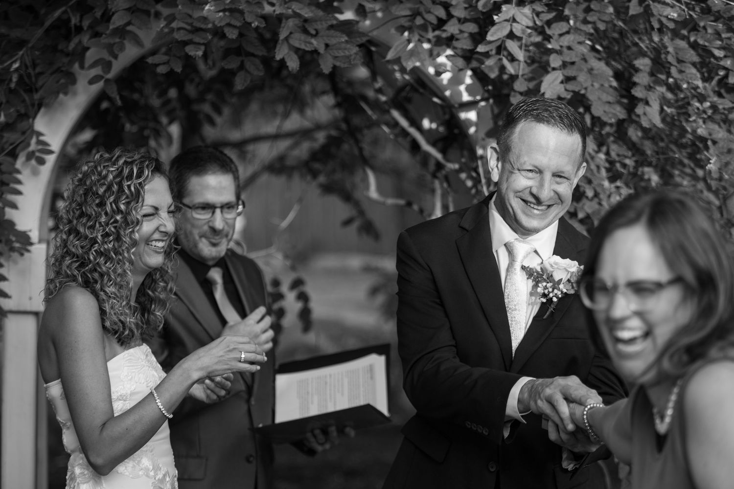Black and white photo of bride and groom laughing with wedding officiant during outdoor ceremony.