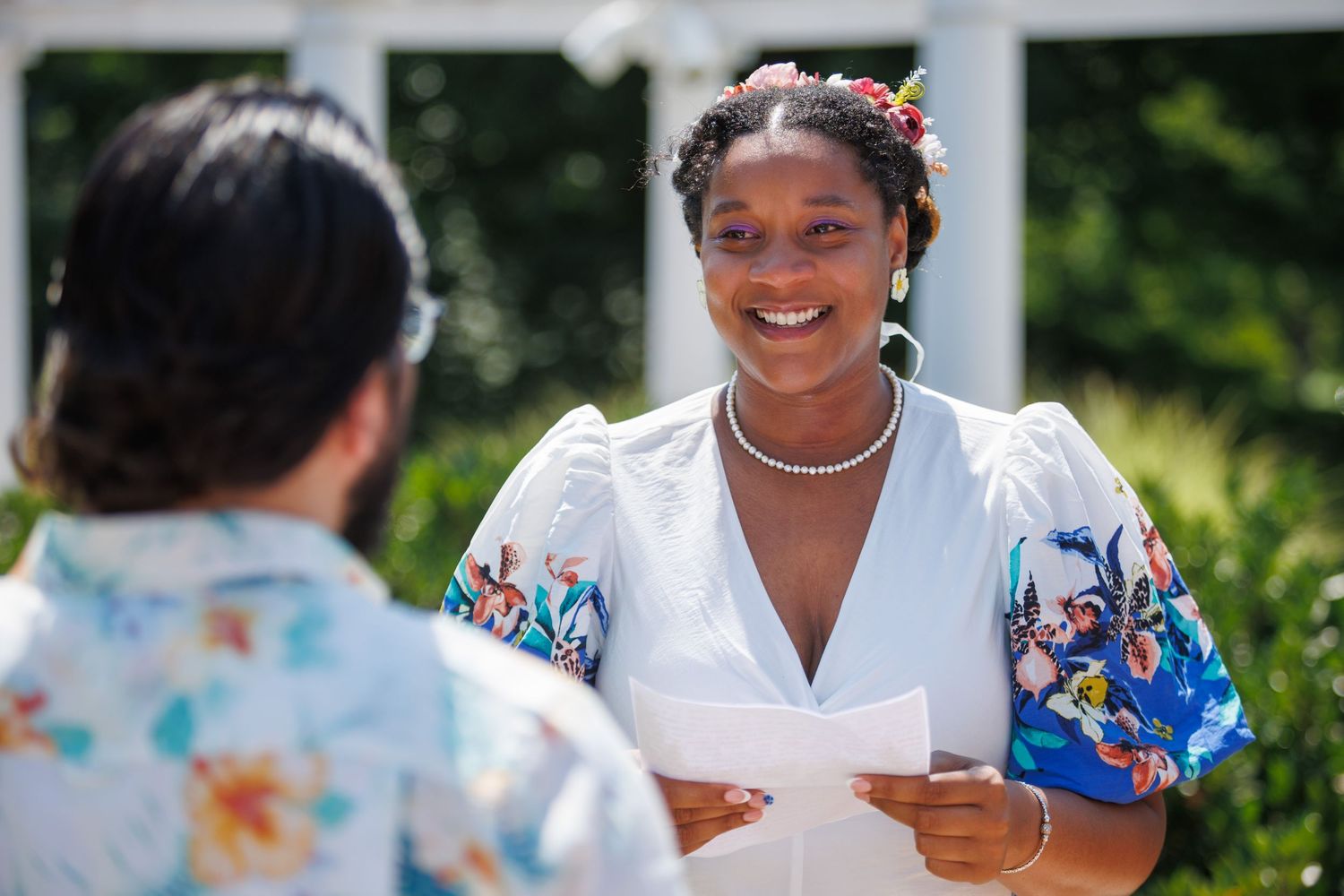 A smiling Black woman in a white floral dress with pearl necklace holds papers outdoors at a sunny garden ceremony.