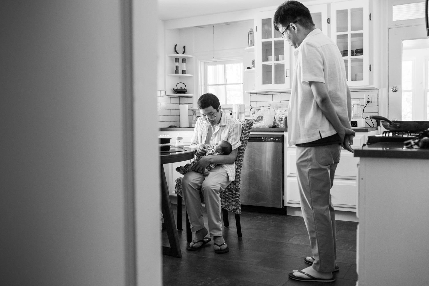 A casual black and white scene in a kitchen with two people standing and sitting while having a conversation.