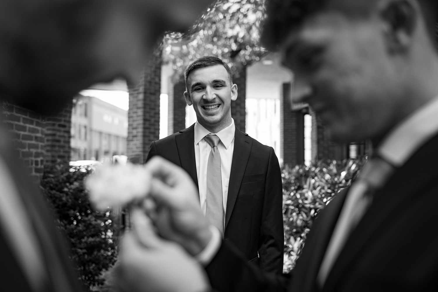 Black and white photo of a smiling groom in a suit and tie chatting with groomsmen outdoors.