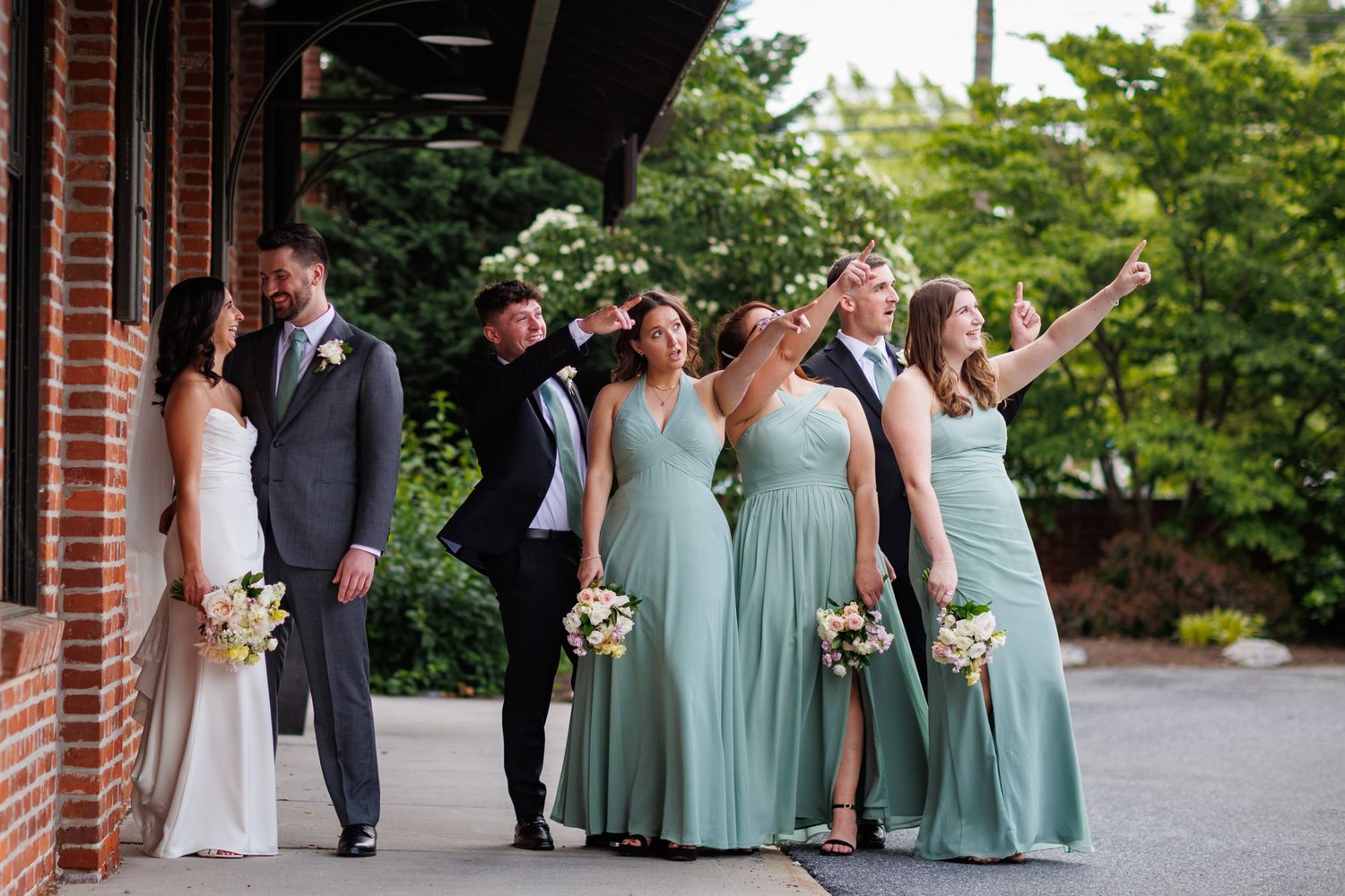 Wedding party in sage green bridesmaid dresses and gray suits posing outdoors near a brick building.