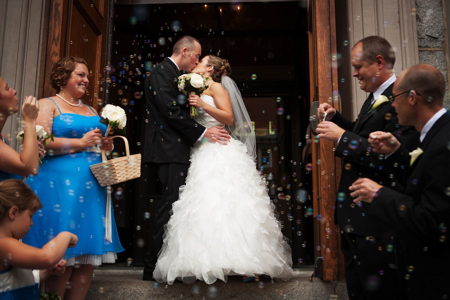 Bride and groom share a kiss at church exit as guests blow bubbles, with bridesmaid in blue dress holding flower basket.