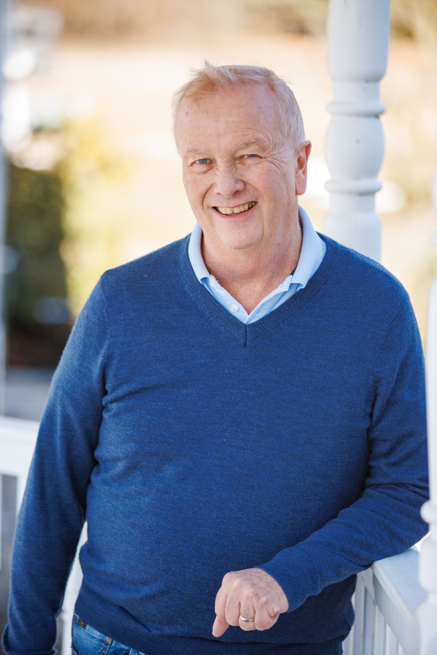 Person wearing a blue sweater smiles warmly while sitting outdoors on a sunny day.