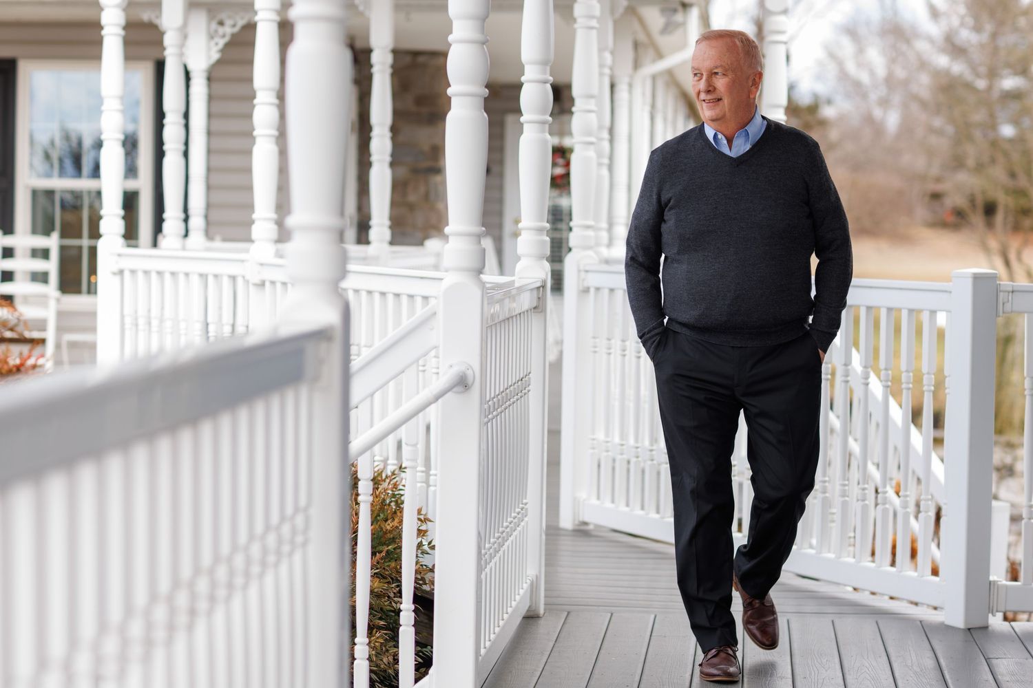 A person in a dark sweater stands on a white wraparound porch with classic columns and railing.