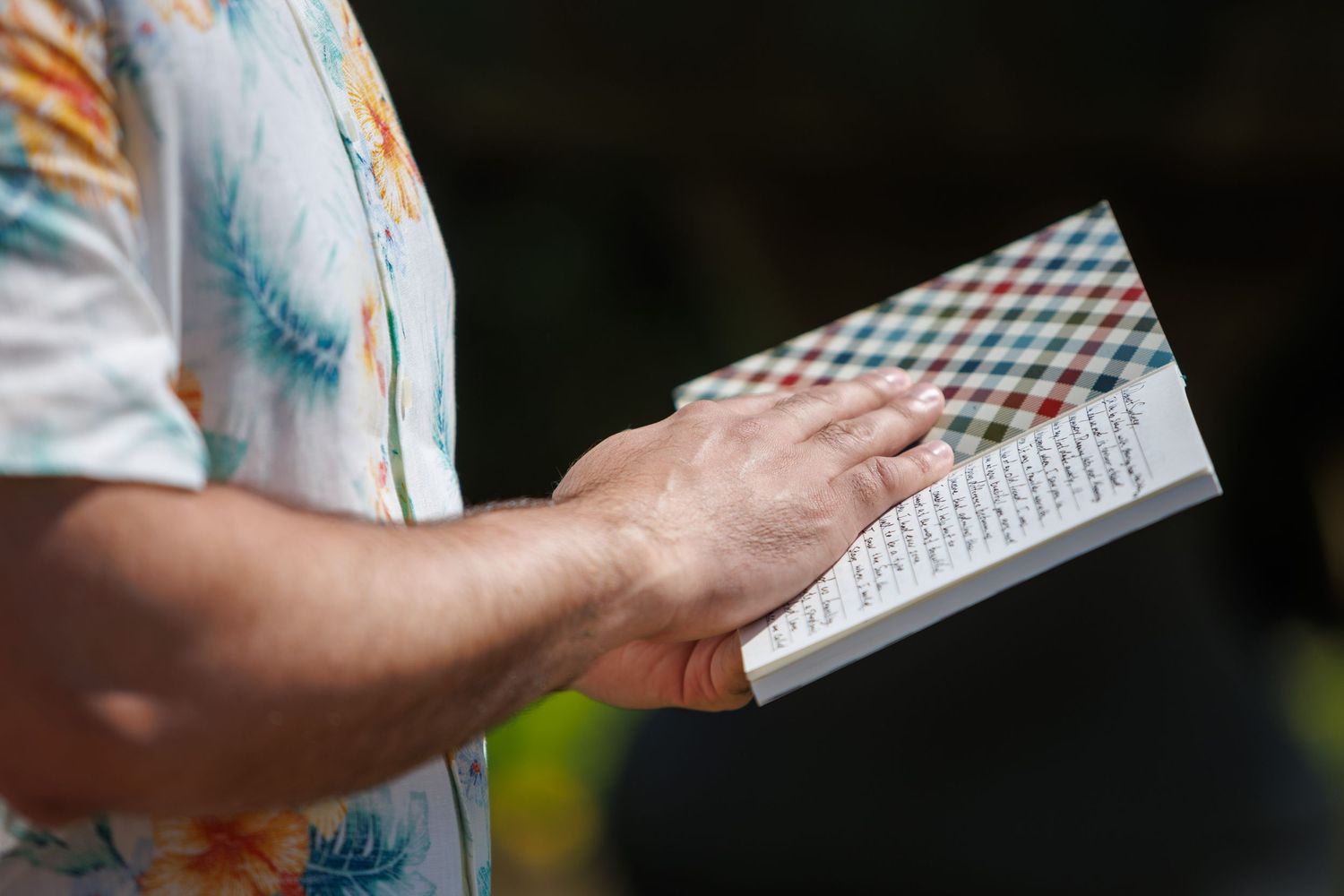 A person in a floral shirt holds an open book with a colorful checkered cover outdoors in sunlight.