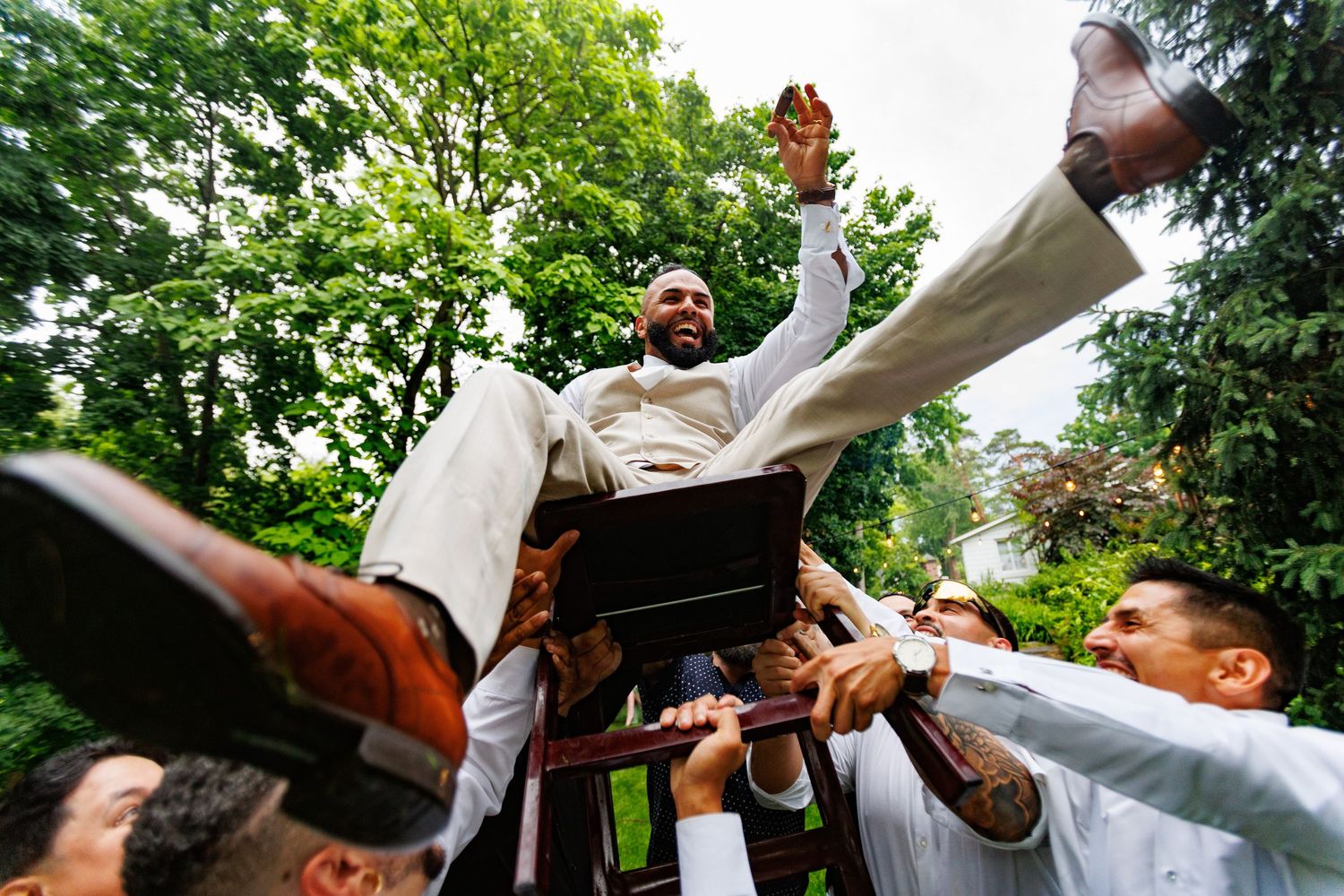 Groom lifted by groomsmen in white suits celebrating joyfully outdoors after wedding ceremony.