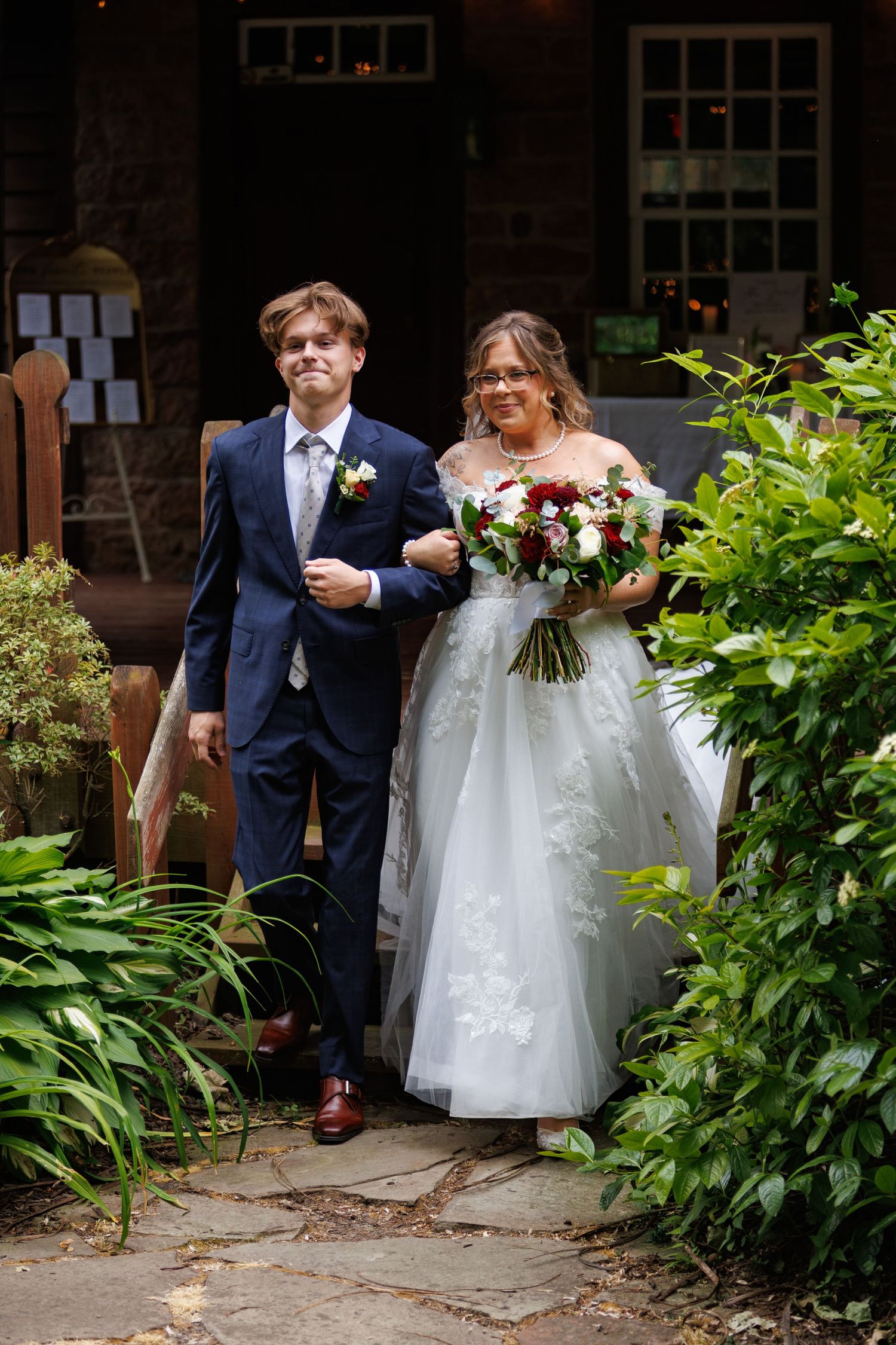 Bride in white lace gown holding red and white bouquet walks with groom in navy suit outdoors.