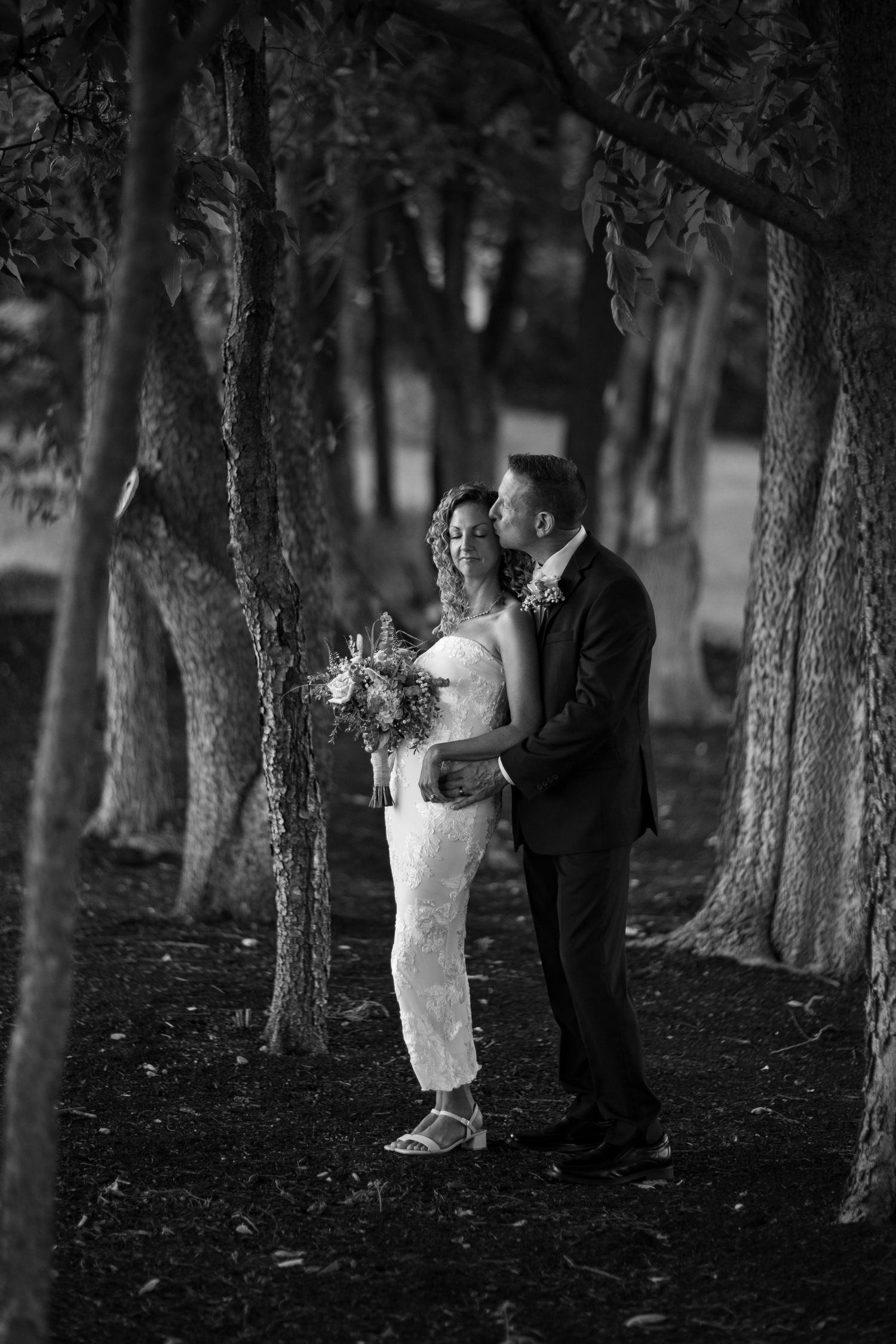Black and white photo of bride and groom embracing among trees in a romantic outdoor forest wedding portrait.
