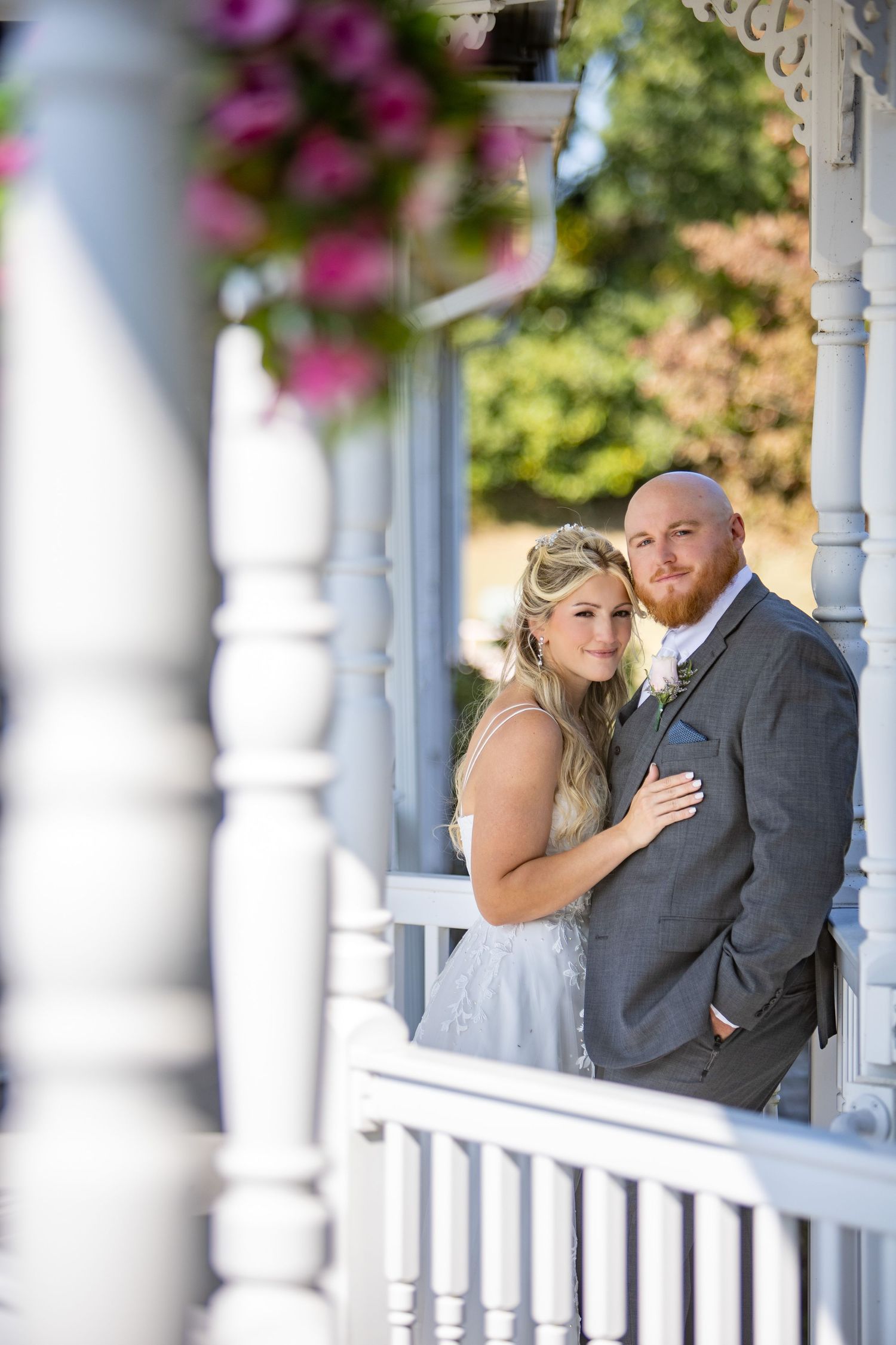 A romantic couple poses together on a white porch with decorative columns and pink flowers in the foreground.