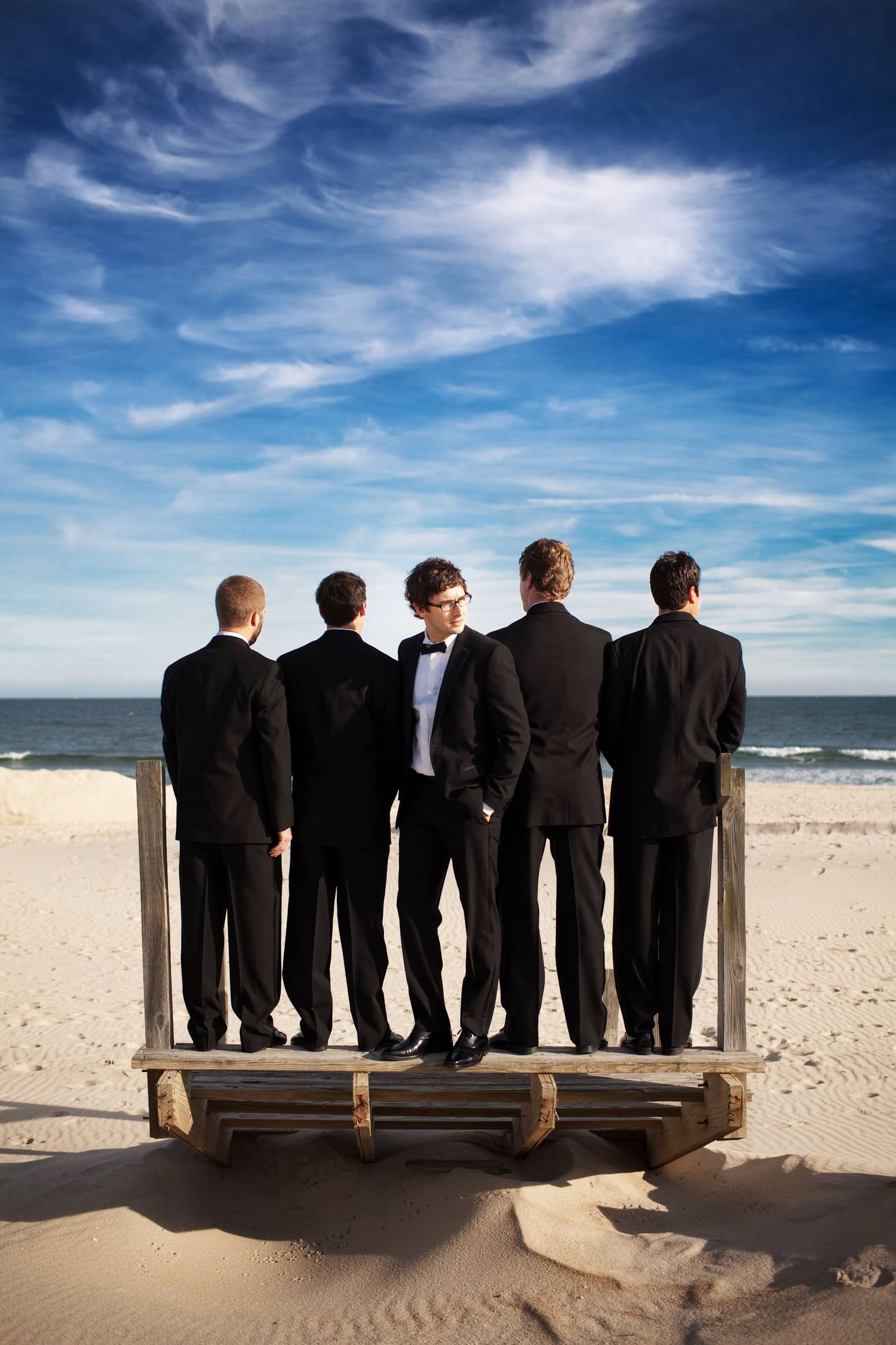 Five groomsmen in black tuxedos standing on a wooden platform at the beach.