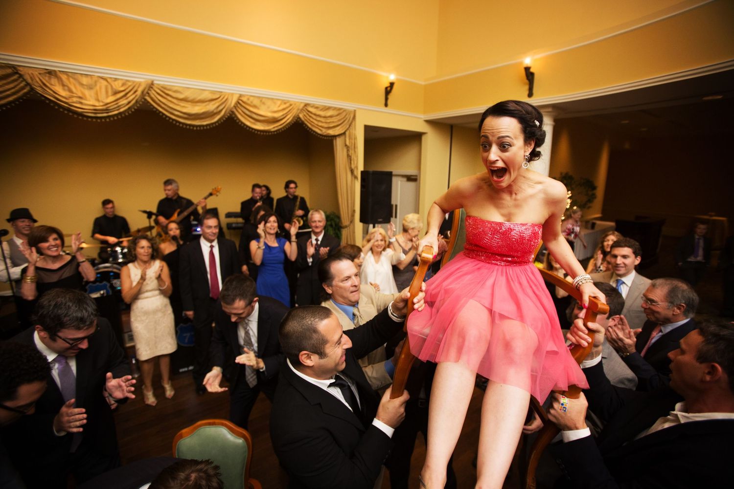 People at a formal event cheer and celebrate as someone in a pink dress dances on a tabletop.