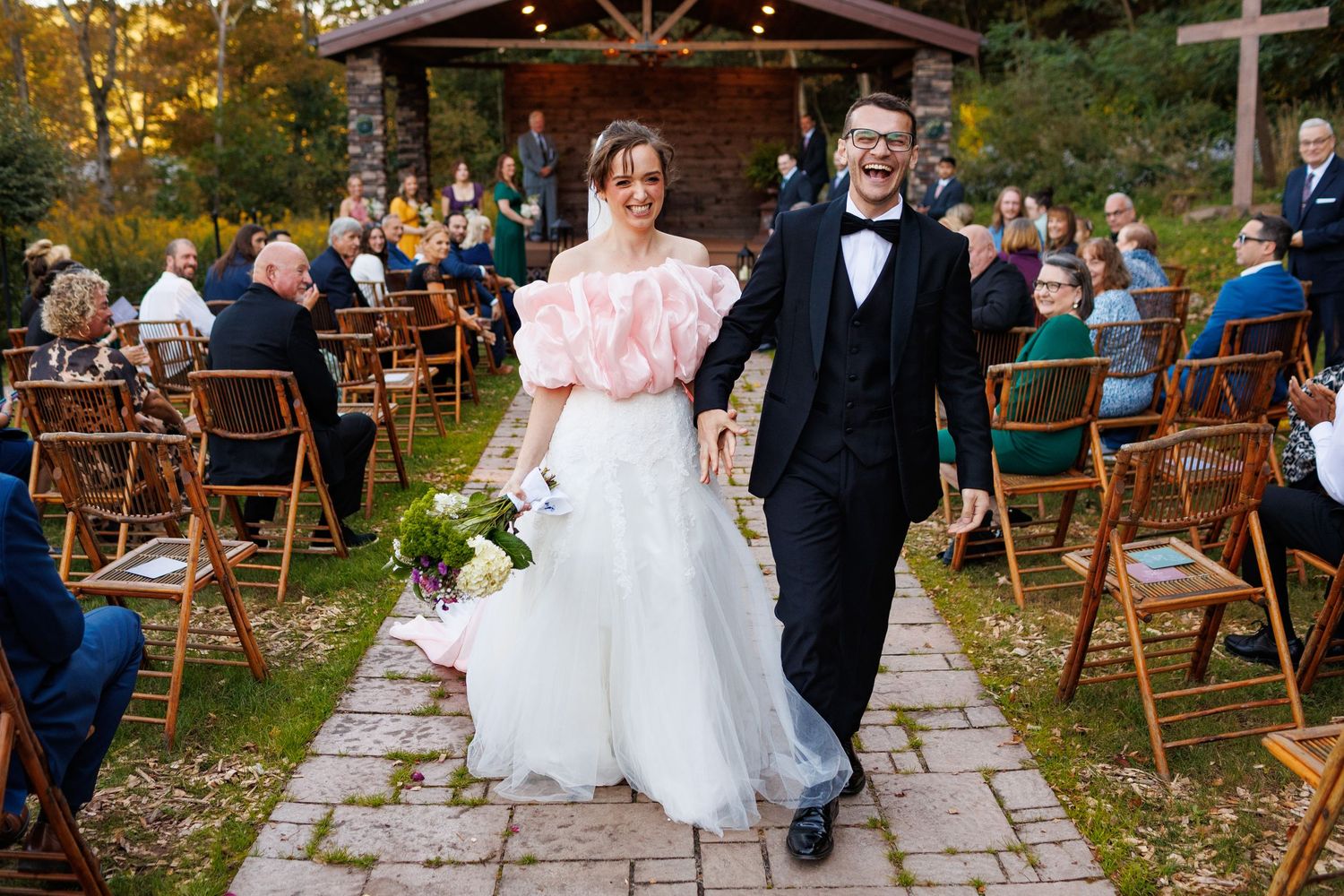 Smiling couple walks up outdoor wedding aisle surrounded by seated guests in autumn forest setting.