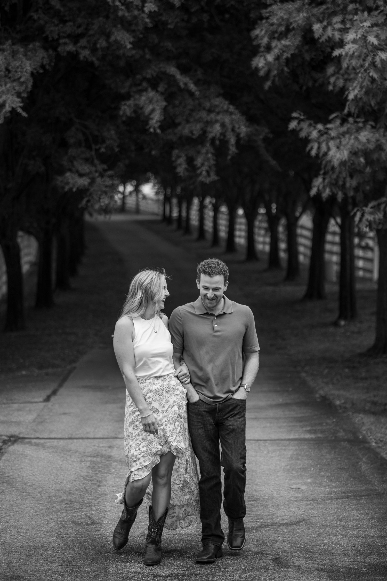Black and white photo of a couple walking hand in hand down a tree-lined path, smiling at each other.