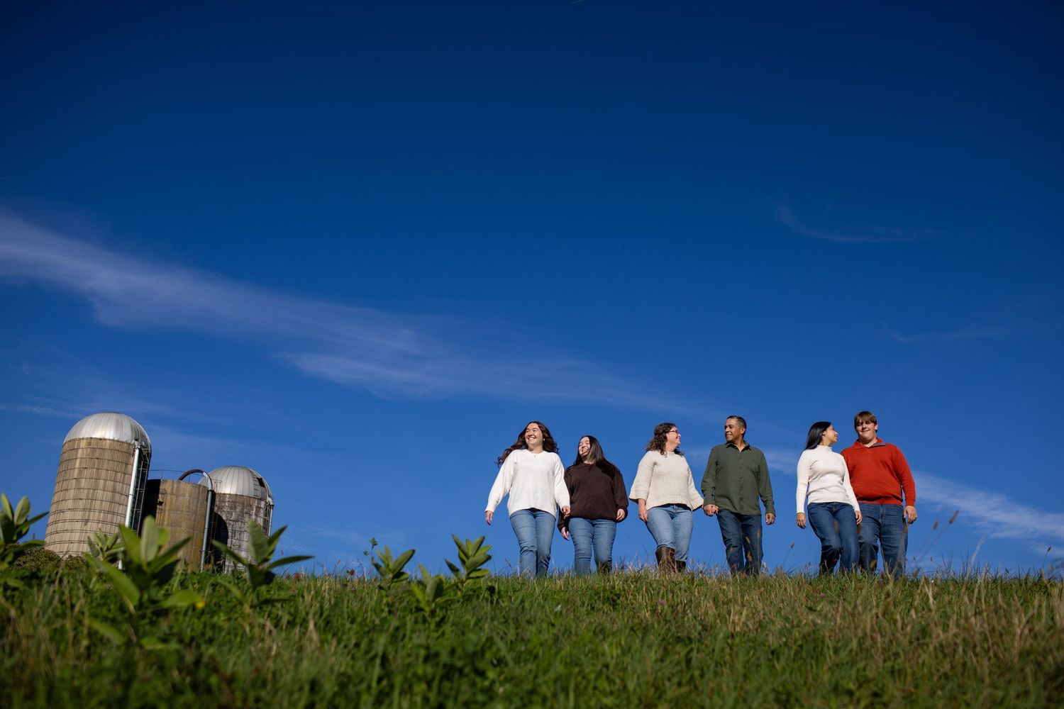 A group of people stand silhouetted against a bright blue sky on a grassy hilltop with buildings in the background.