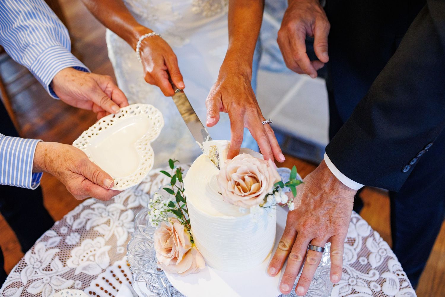 Bride and groom cutting elegant white wedding cake decorated with roses on a lace tablecloth.