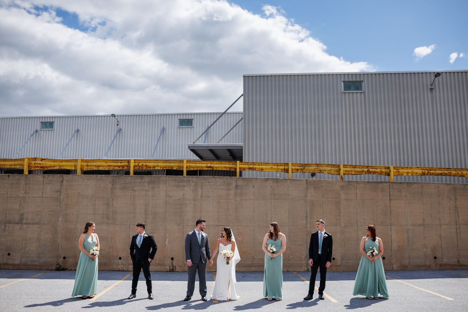 Wedding party poses outdoors in sage green bridesmaid dresses and suits against an industrial building backdrop.