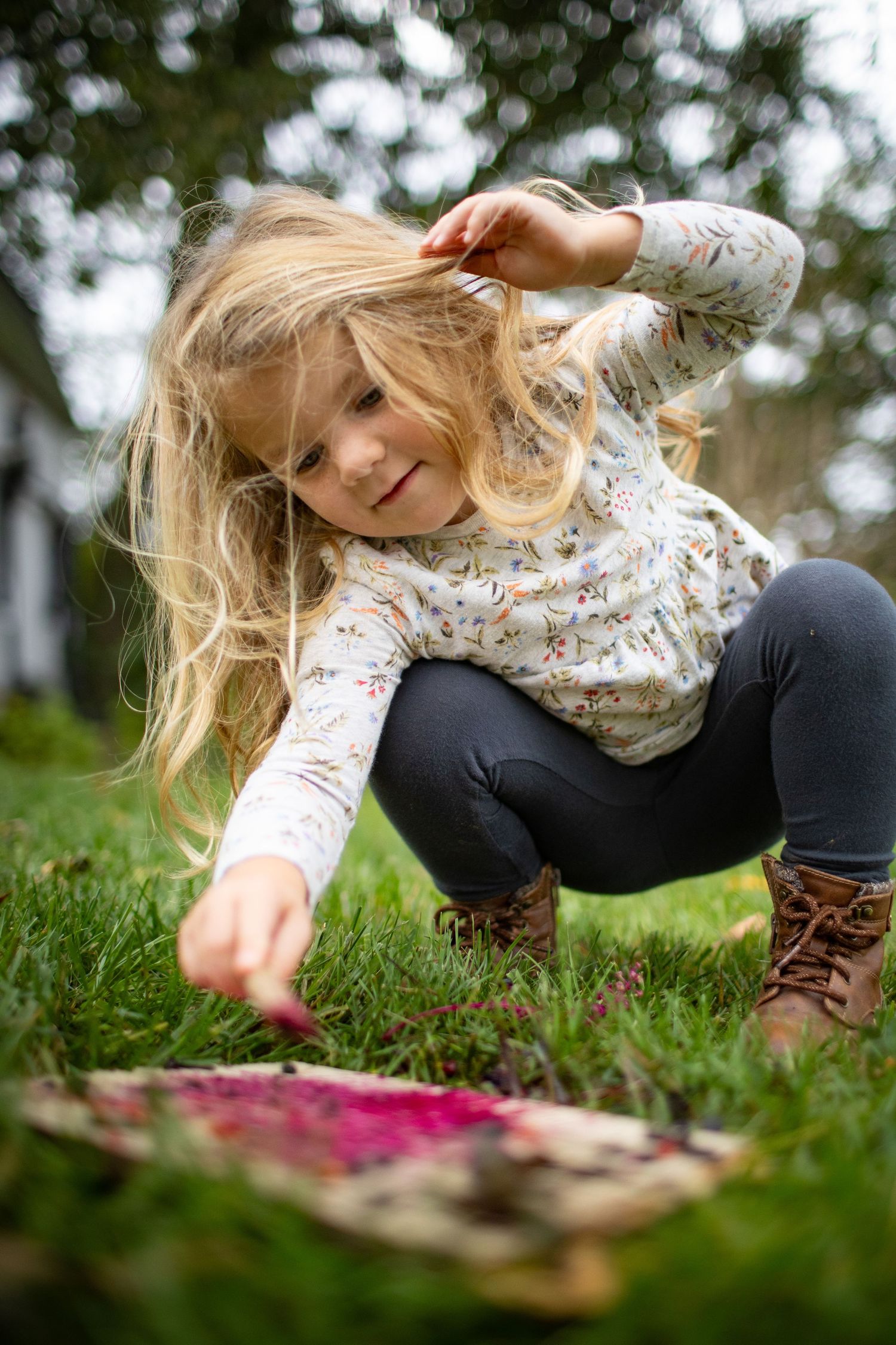 A person in brown boots and black pants reaches down to touch pink flowers growing in green grass.