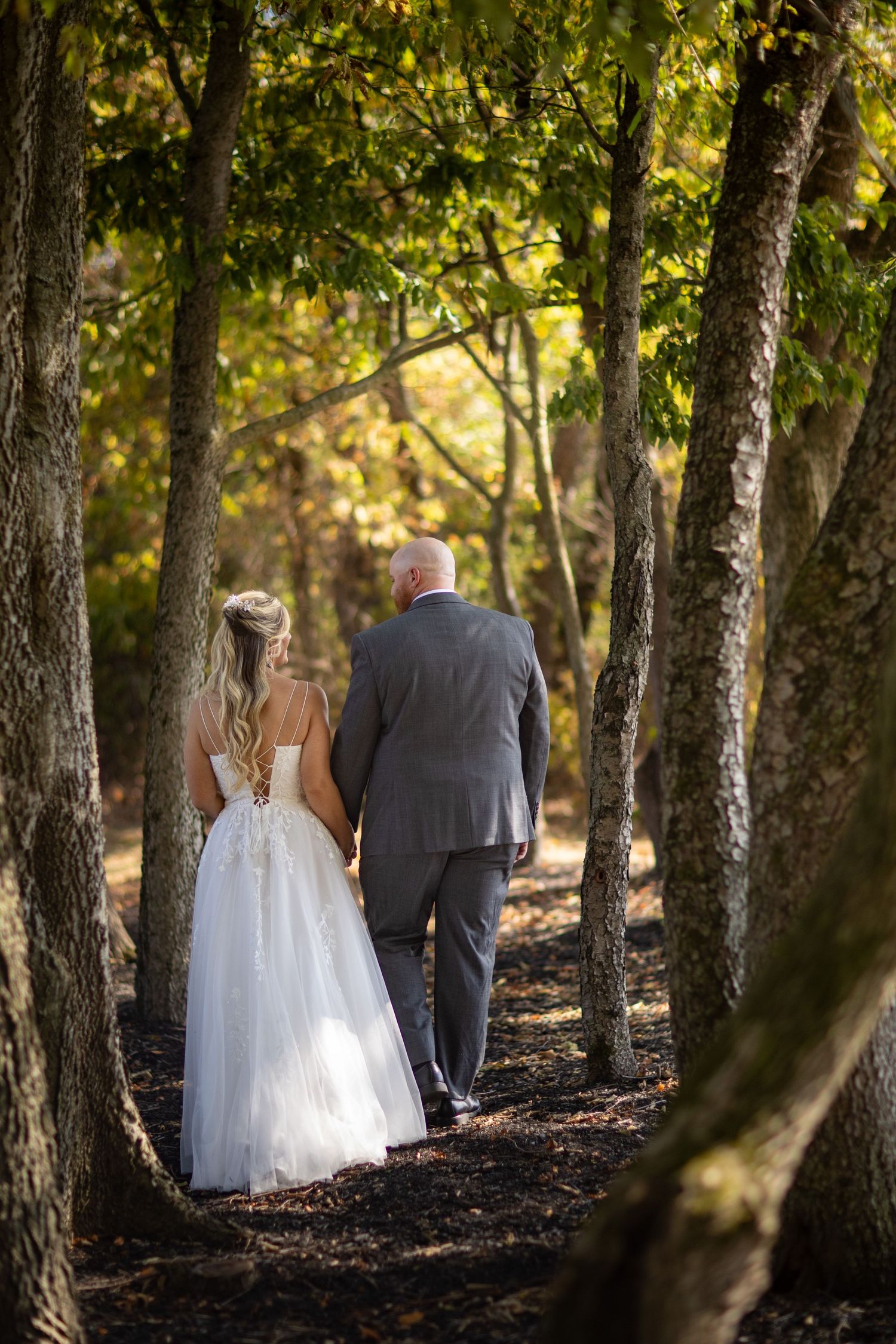A couple in wedding attire walking down a tree-lined woodland path in dappled sunlight during autumn.