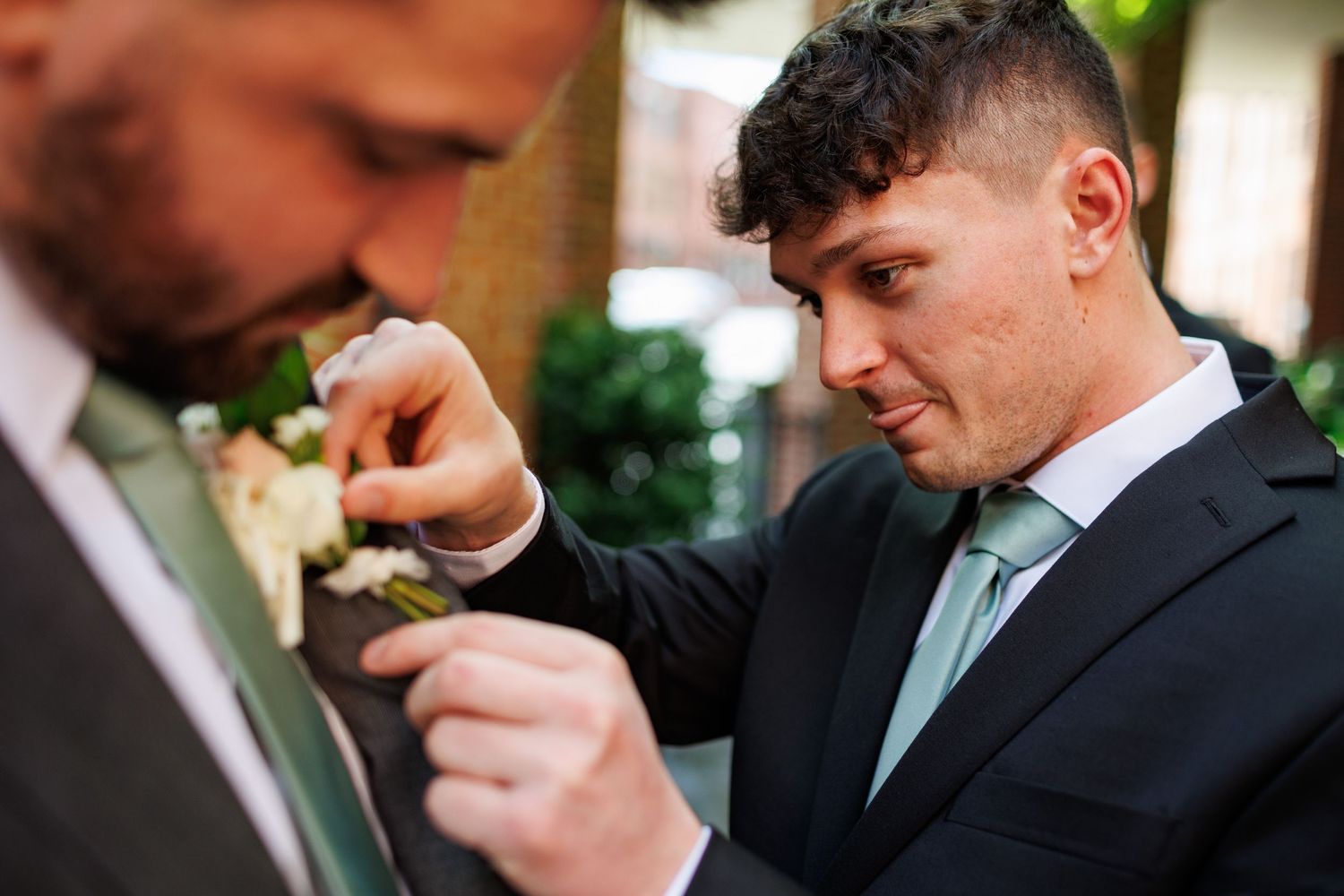 Groomsman carefully pins a white boutonniere onto the lapel of a groom in a dark suit with green tie.