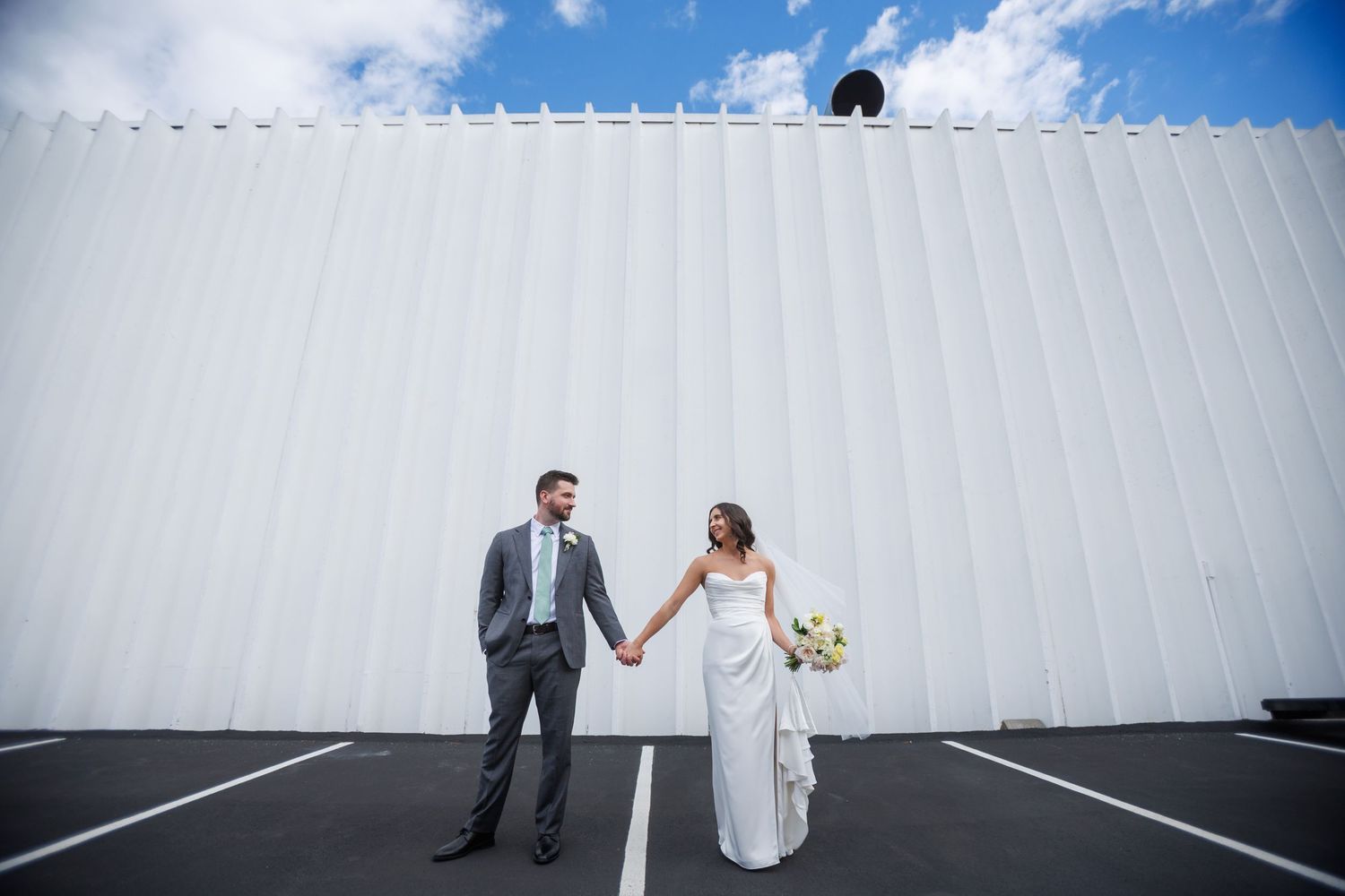 Bride and groom holding hands in a parking lot against a white building under a bright blue sky.