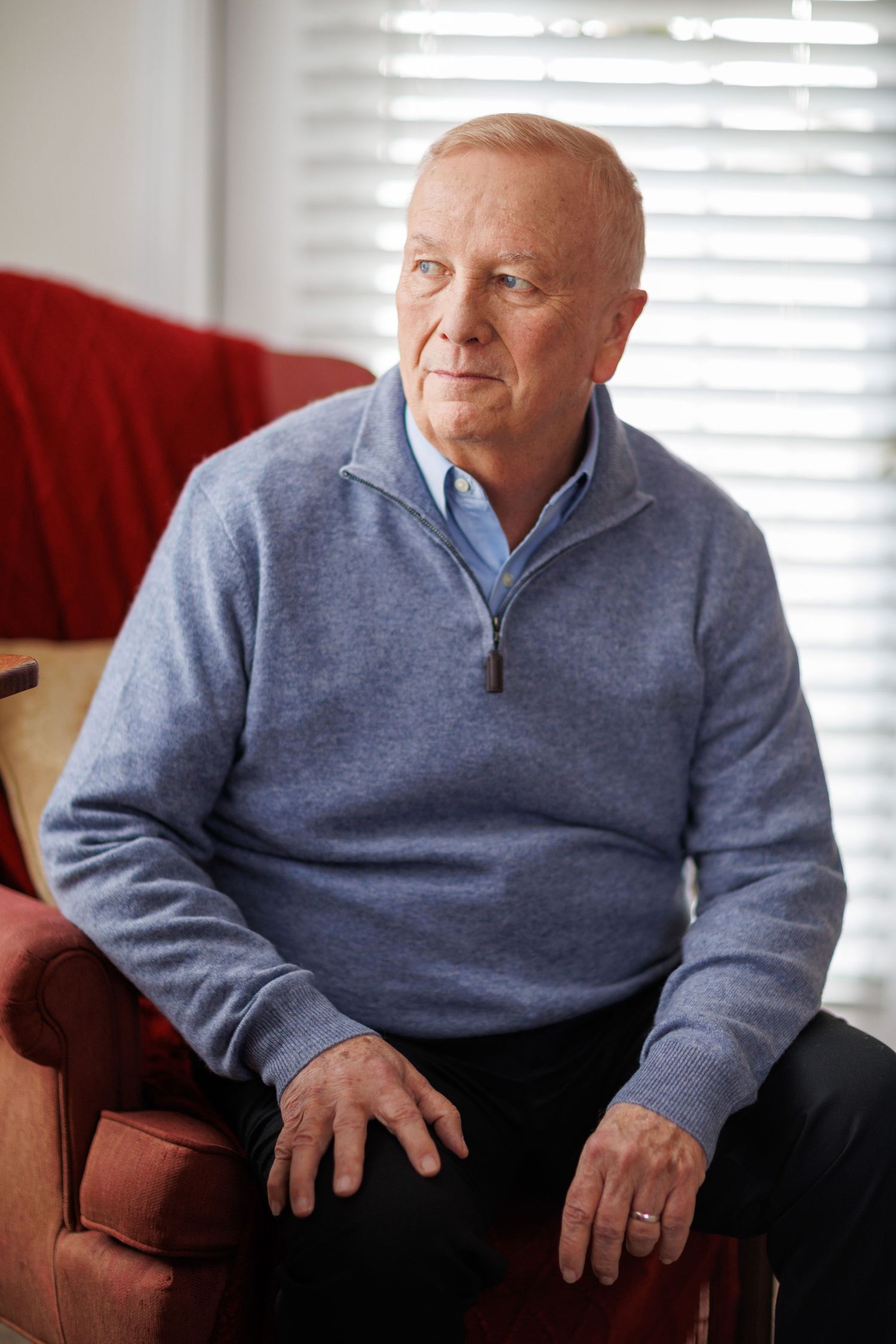 Senior man in blue quarter-zip sweater sits thoughtfully on a red armchair near window with white blinds.