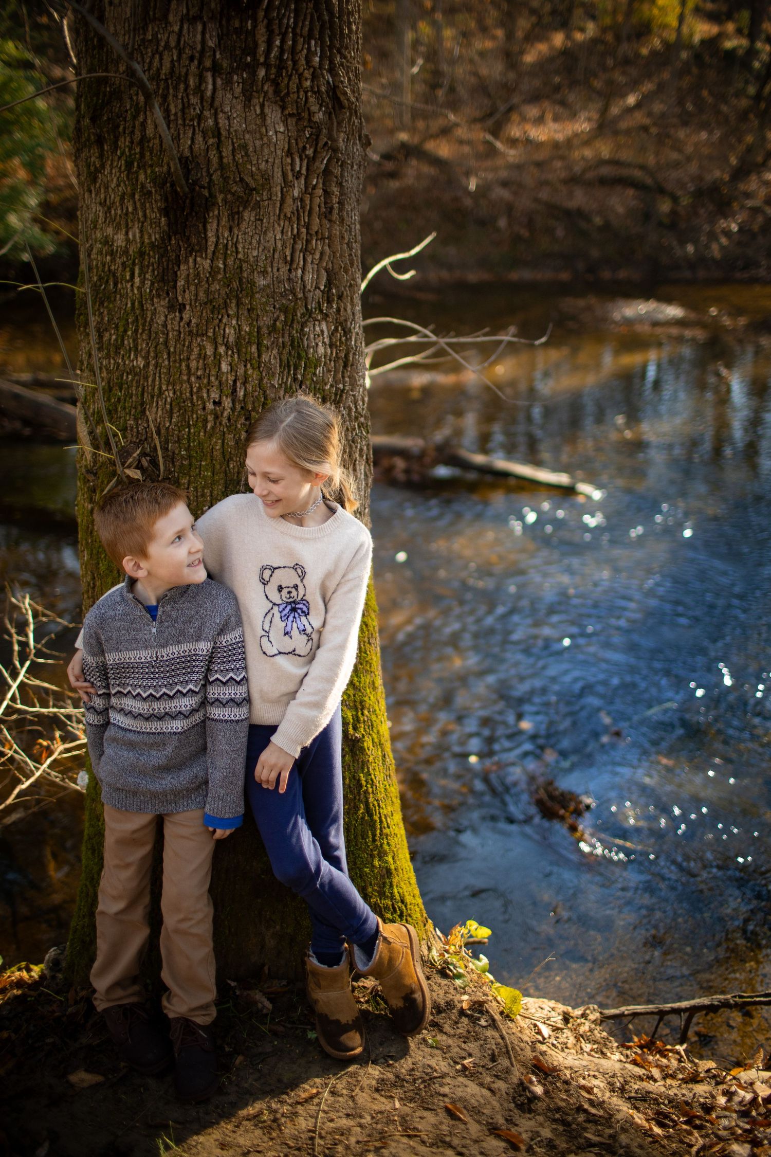 People sitting together by a tree trunk near a reflective stream during fall season.