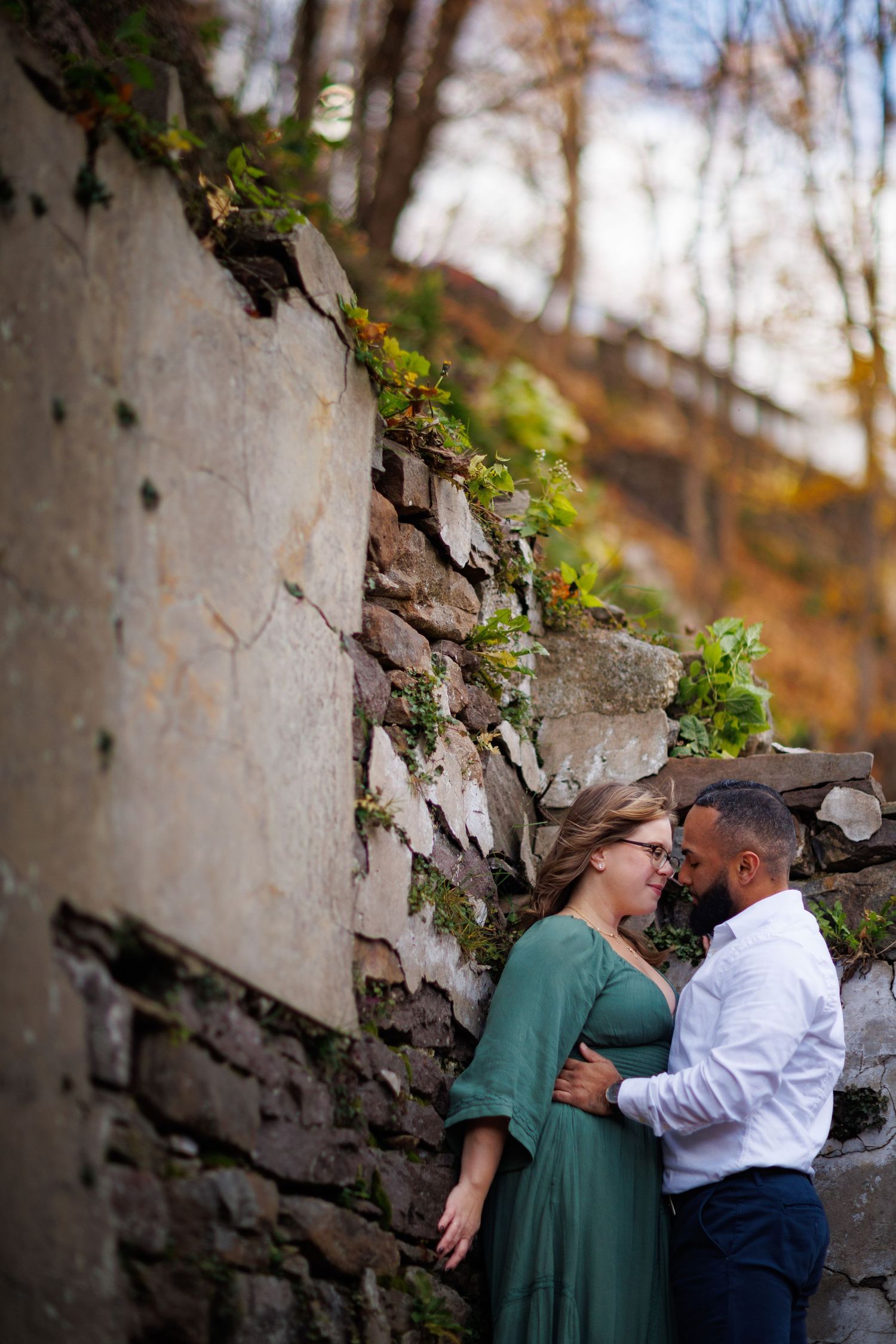 A romantic engaged couple embraces against a stone wall backdrop during an autumn outdoor photoshoot.